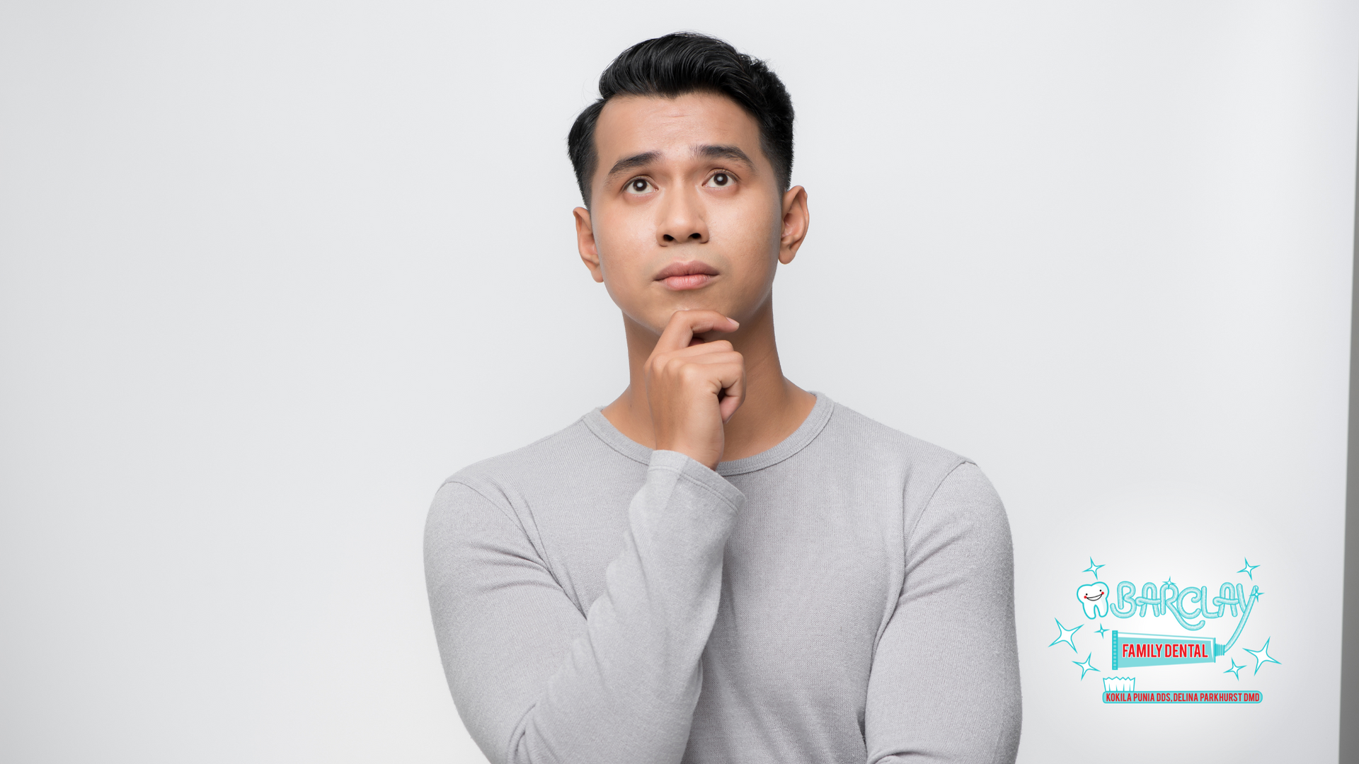 Asian man in gray sweater, gazing upward thoughtfully, hand on chin, white background.