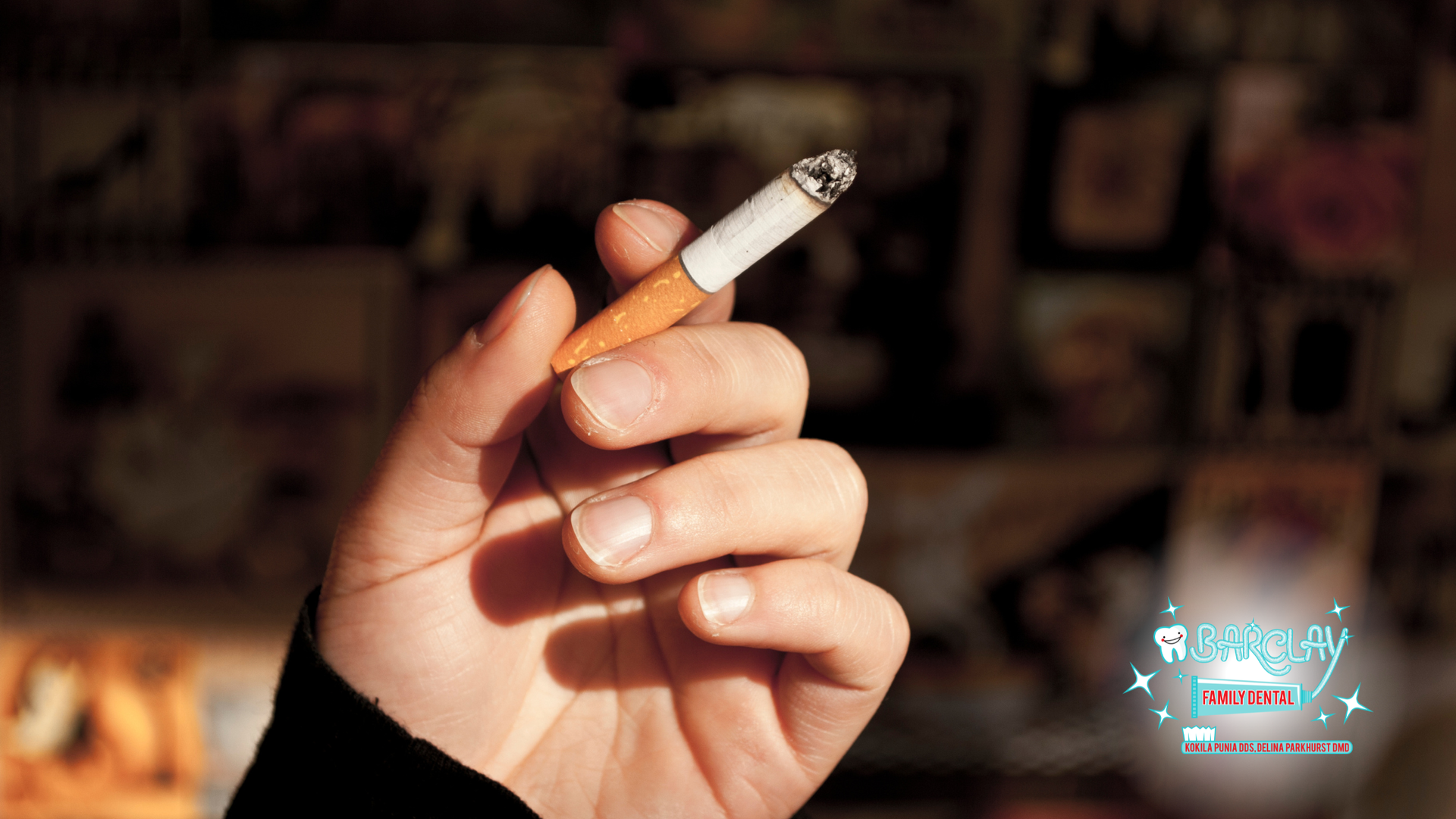 Hand holding a lit cigarette, indoors against a blurry background of photos.