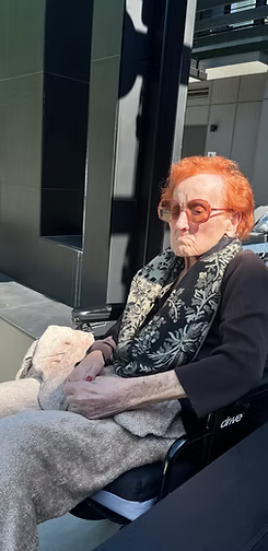 Person with orange hair and sunglasses sitting in a wheelchair indoors, covered with a blanket