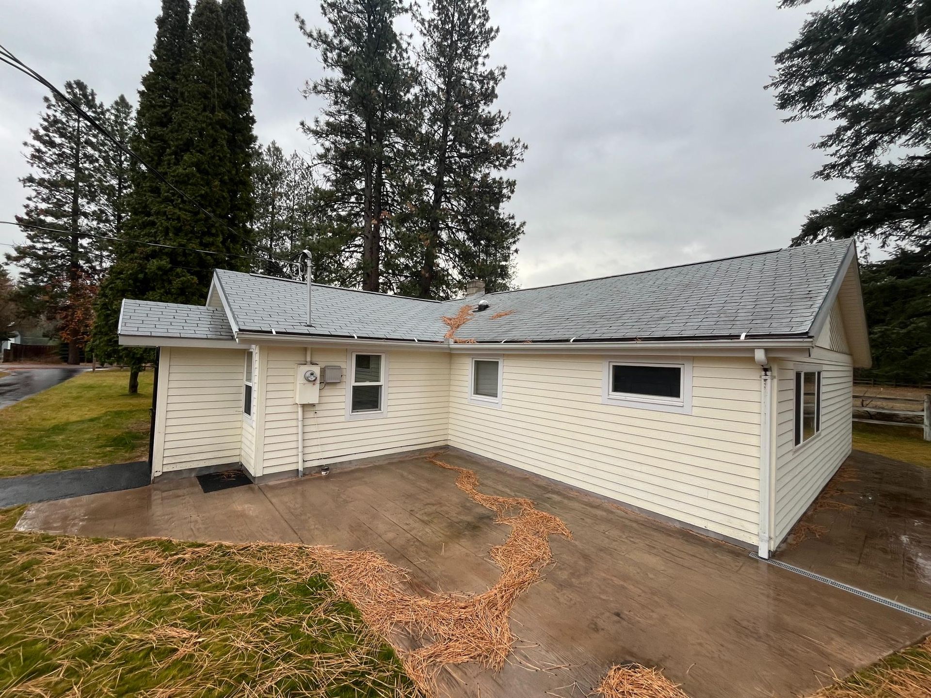 Beige house with gray roof, patio, and fallen leaves on a gray driveway, with green grass and trees in the background.