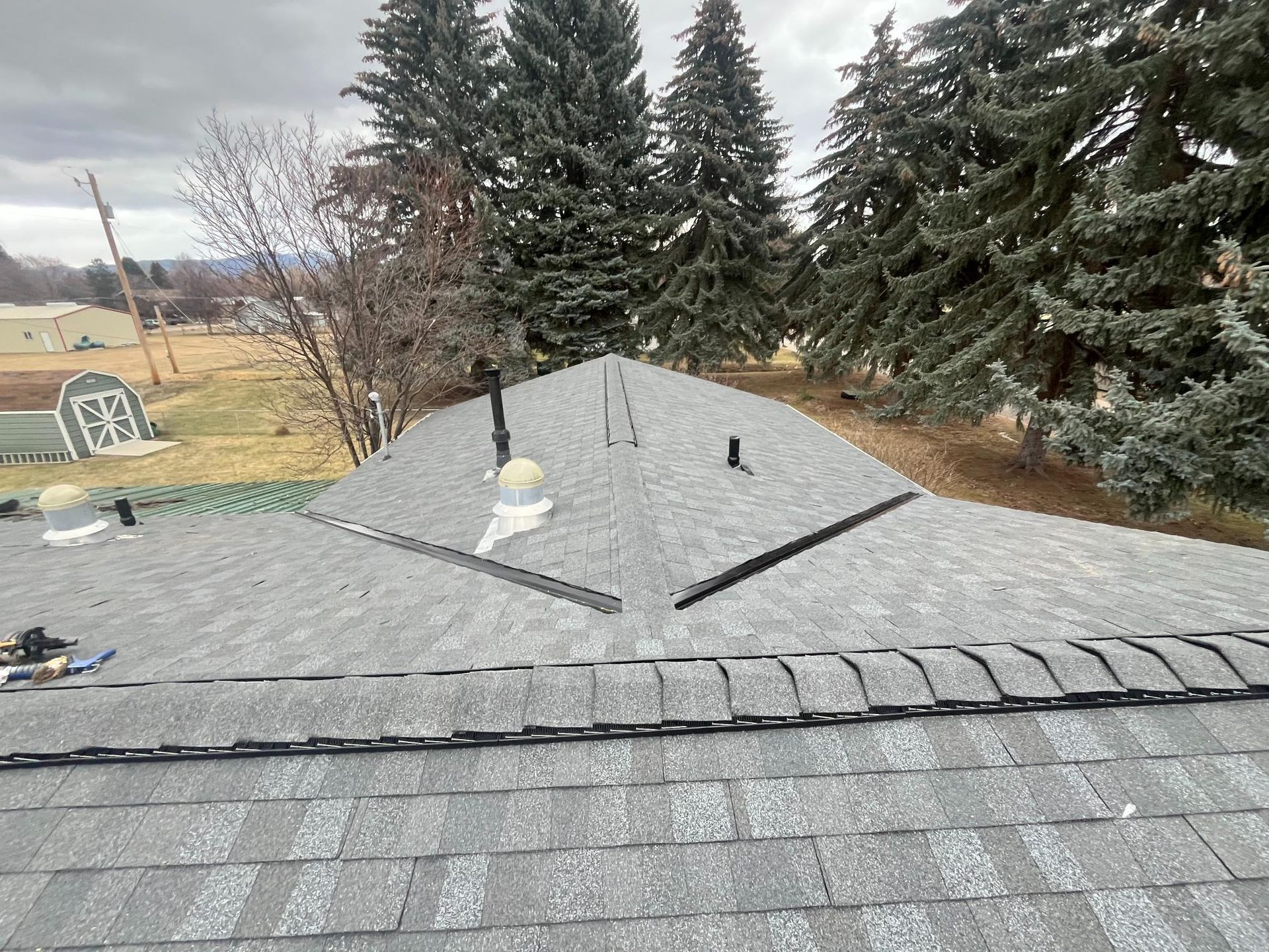 Gray shingle roof with black metal flashing and vents, trees and a distant field visible.