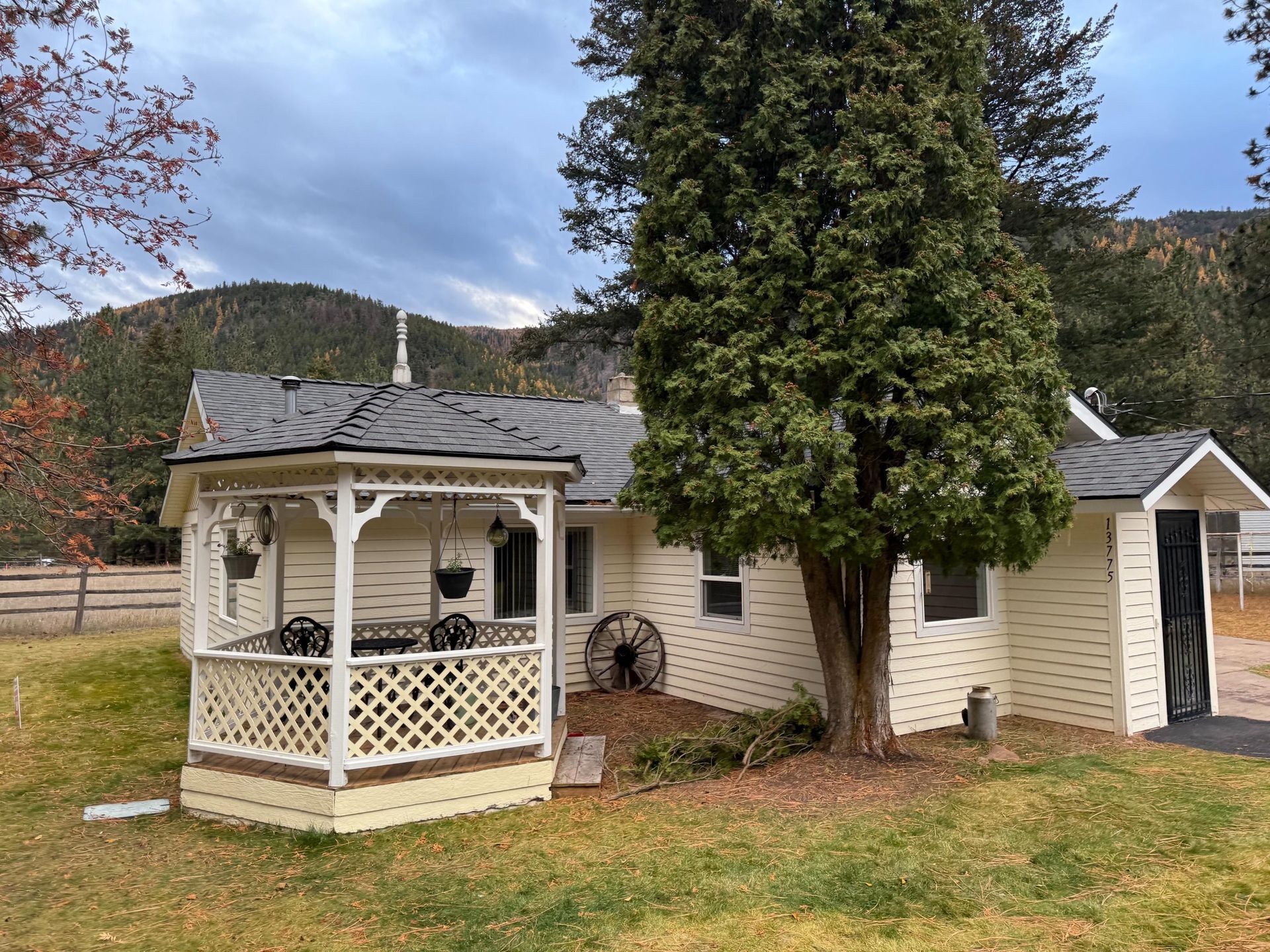 Yellow house with gazebo, surrounded by trees and grass, mountains in the background.
