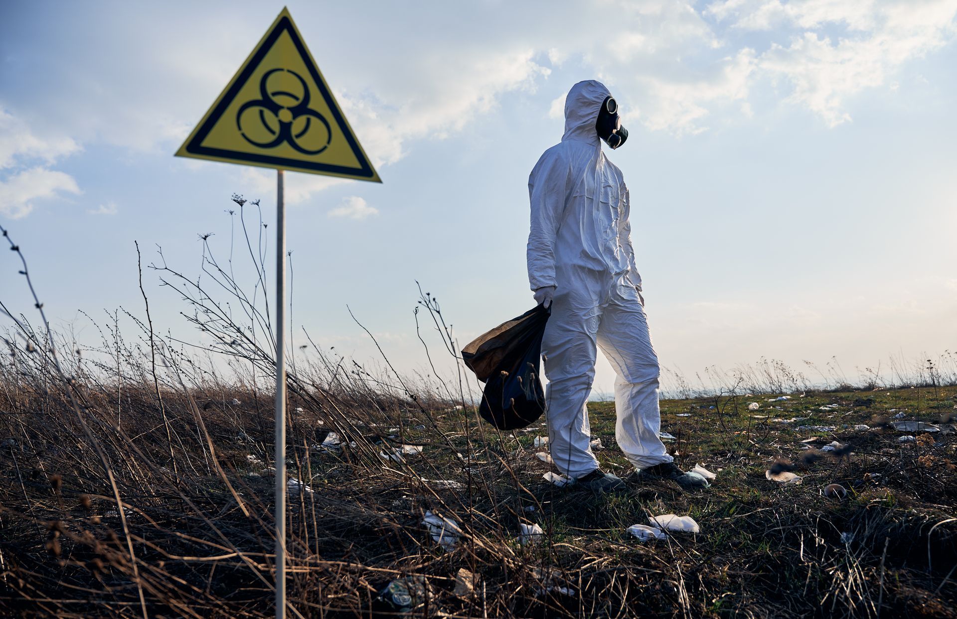Person in hazmat suit stands near a biohazard sign in a contaminated field, under a blue sky.