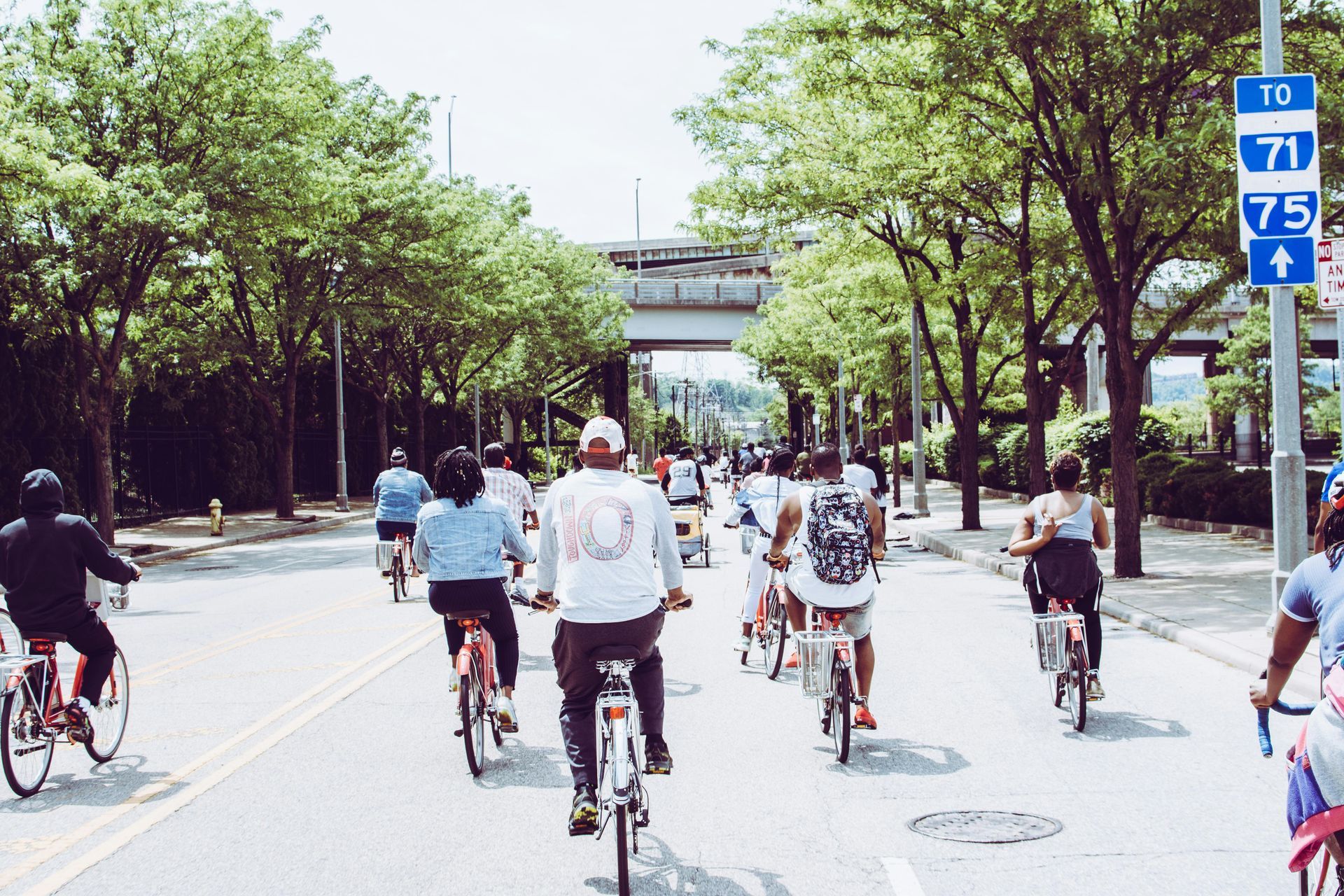 Group of people biking on a sunny street lined with trees. A blue highway sign is visible.