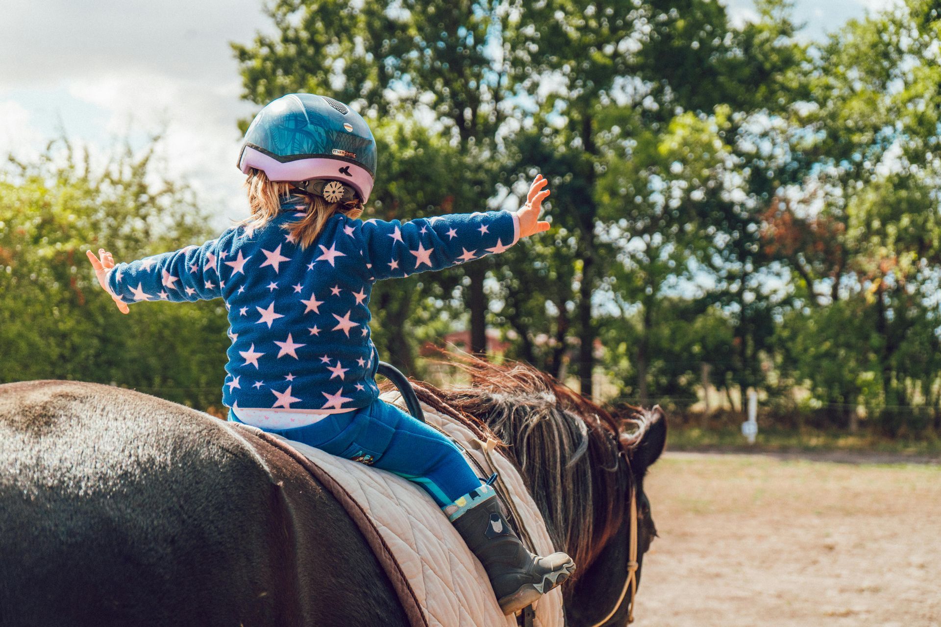 Young child on a horse, wearing a helmet and blue star-patterned sweater, arms outstretched. Outdoors, sunny day.