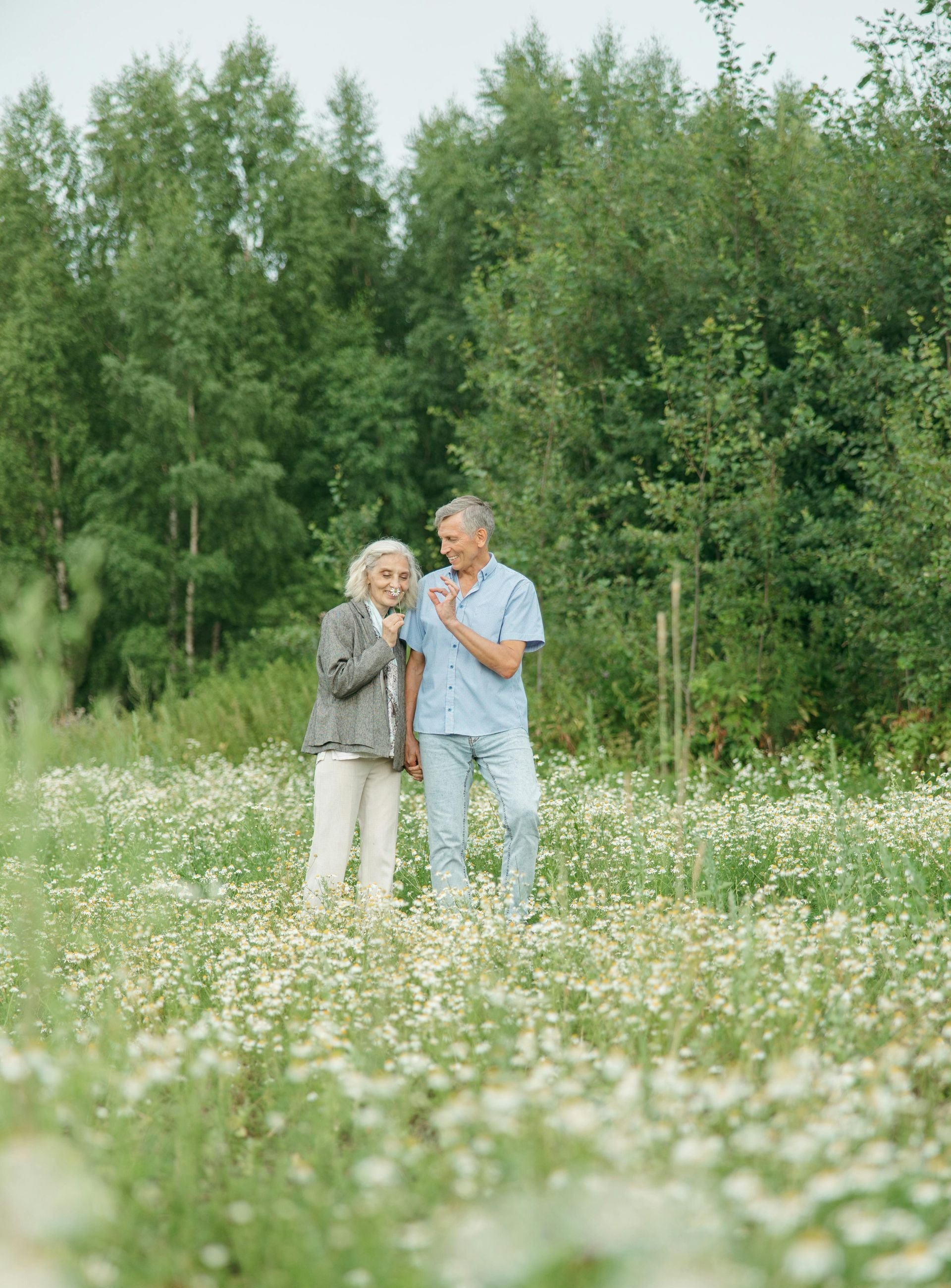 Elderly couple standing in a field of daisies, smiling at the camera. Green trees in the background.