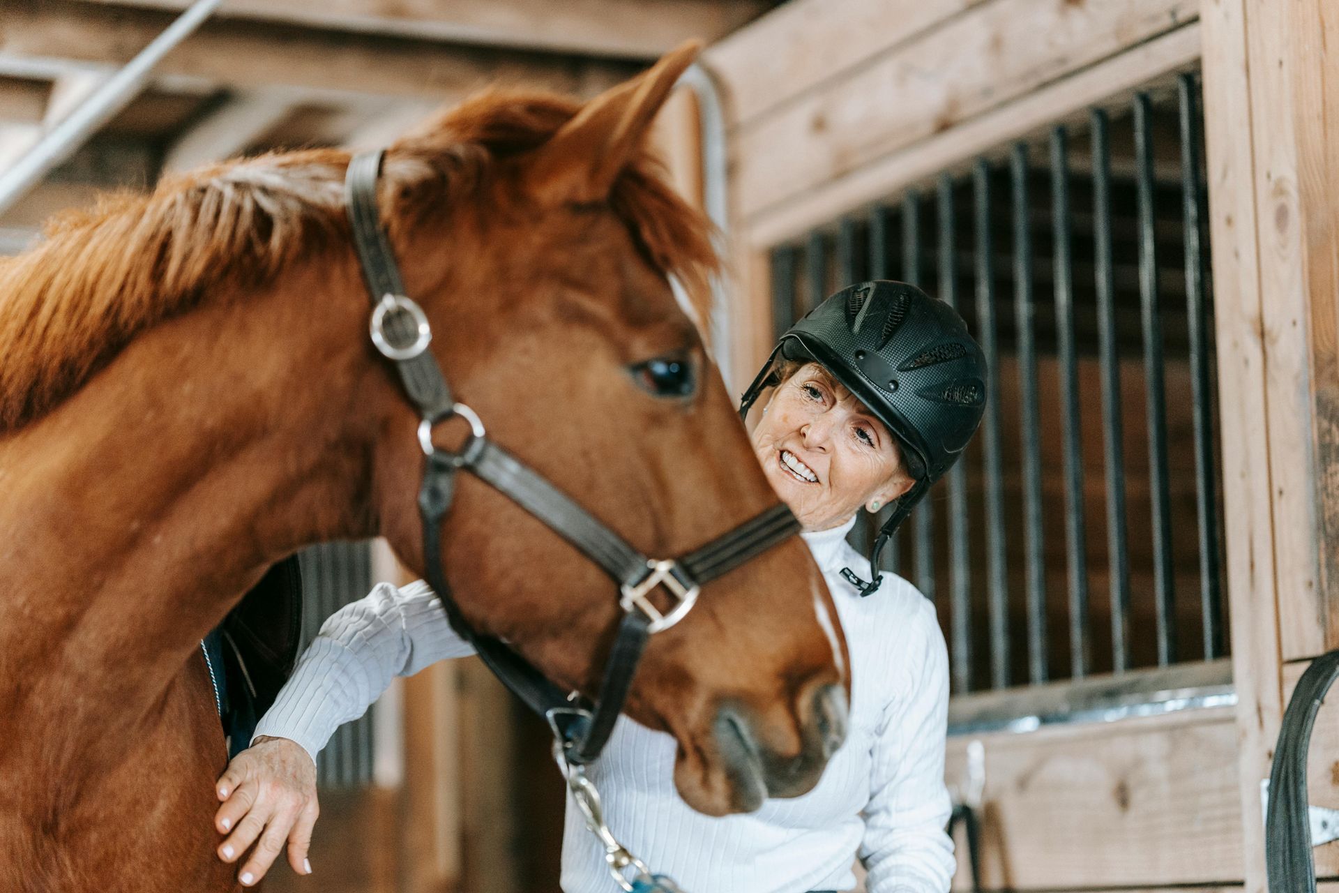 Woman in riding gear smiles, resting against a brown horse in a stable.