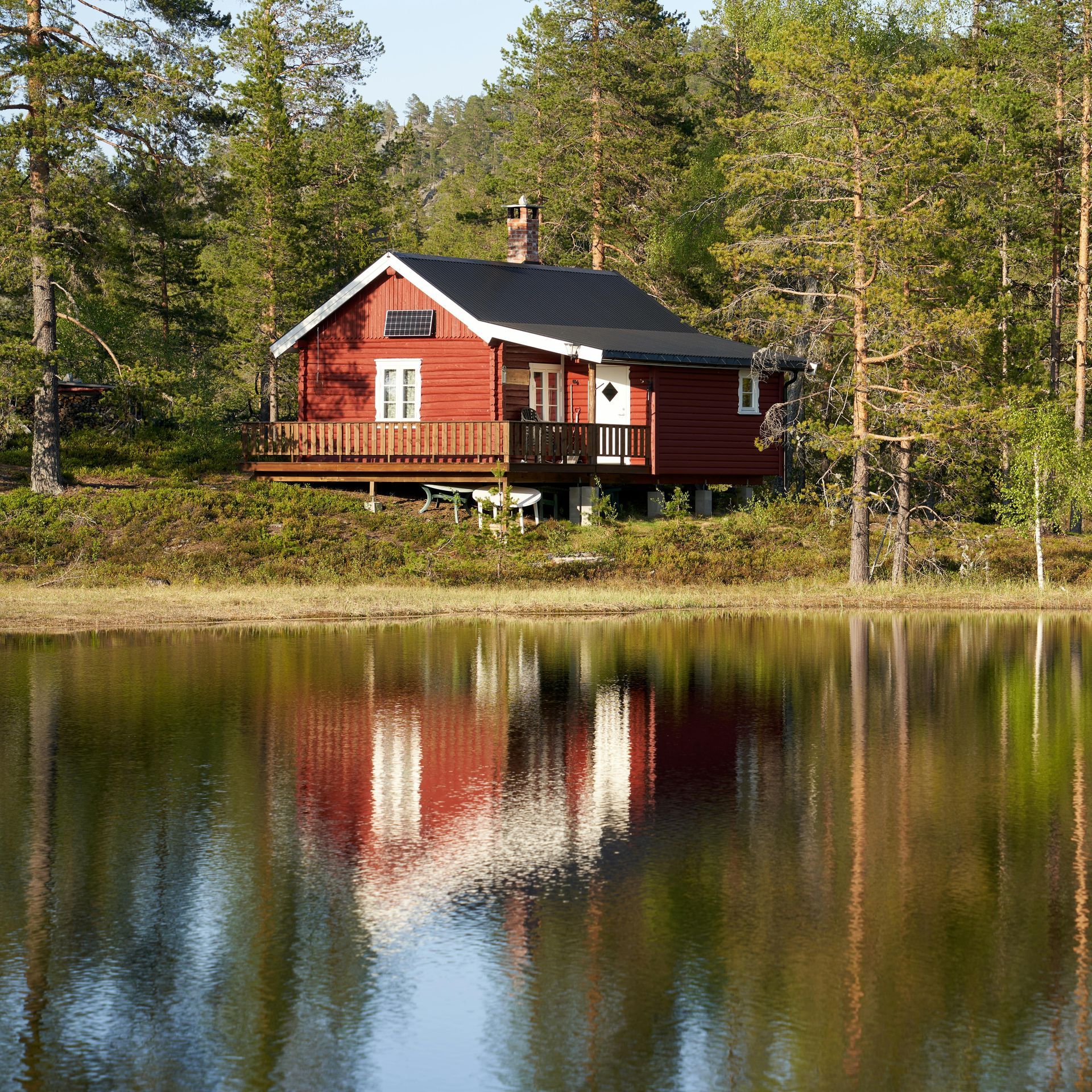Red cabin with a black roof sits on a bank reflecting in calm water; surrounded by trees.
