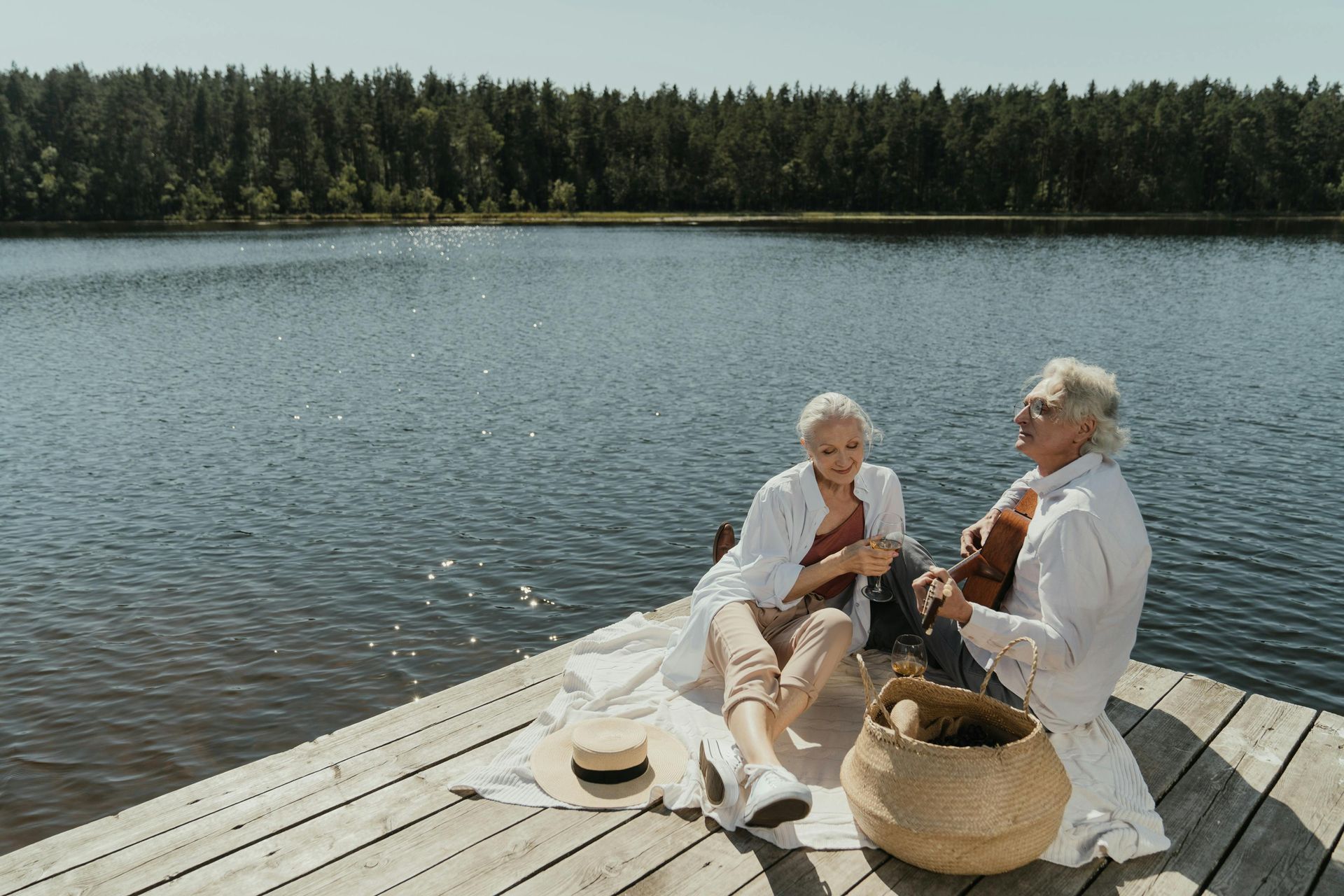 Two older adults sit on a wooden dock by a lake. A woman watches as a man plays a guitar; both are smiling in the sun.