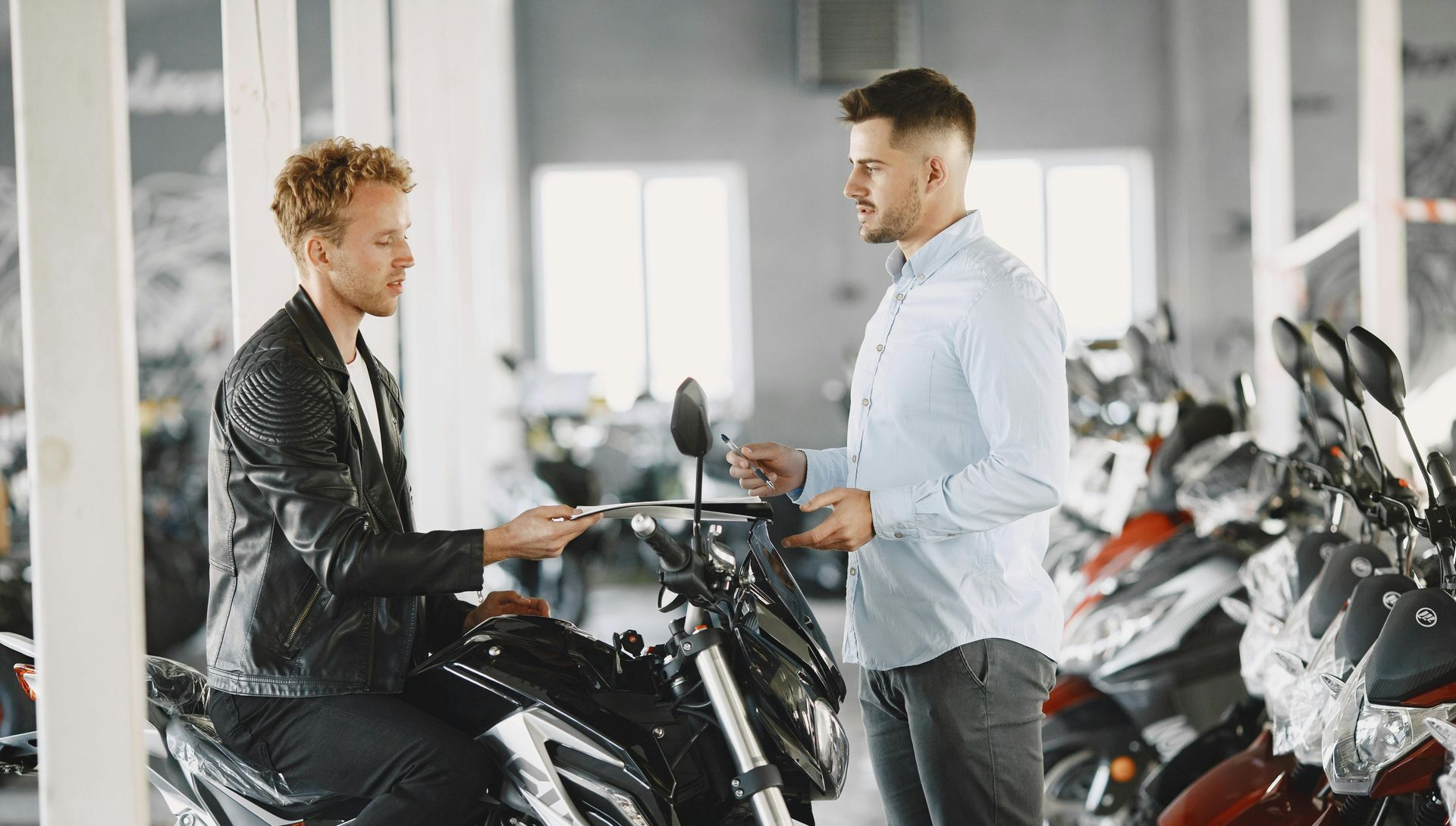 Man in leather jacket on motorcycle receives keys from salesman in showroom.