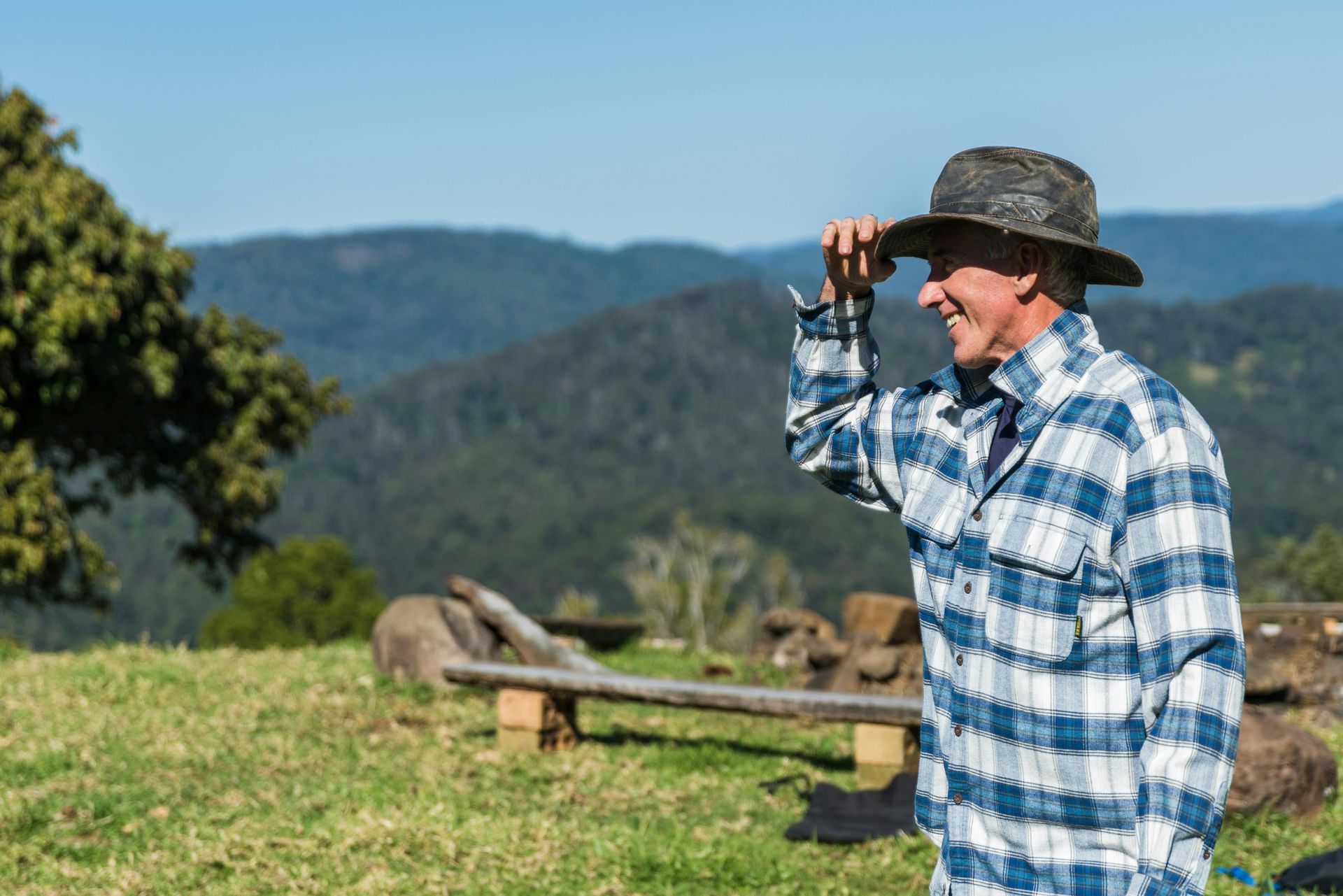 Man in plaid shirt and hat, shielding his eyes and smiling while looking at a mountain range.