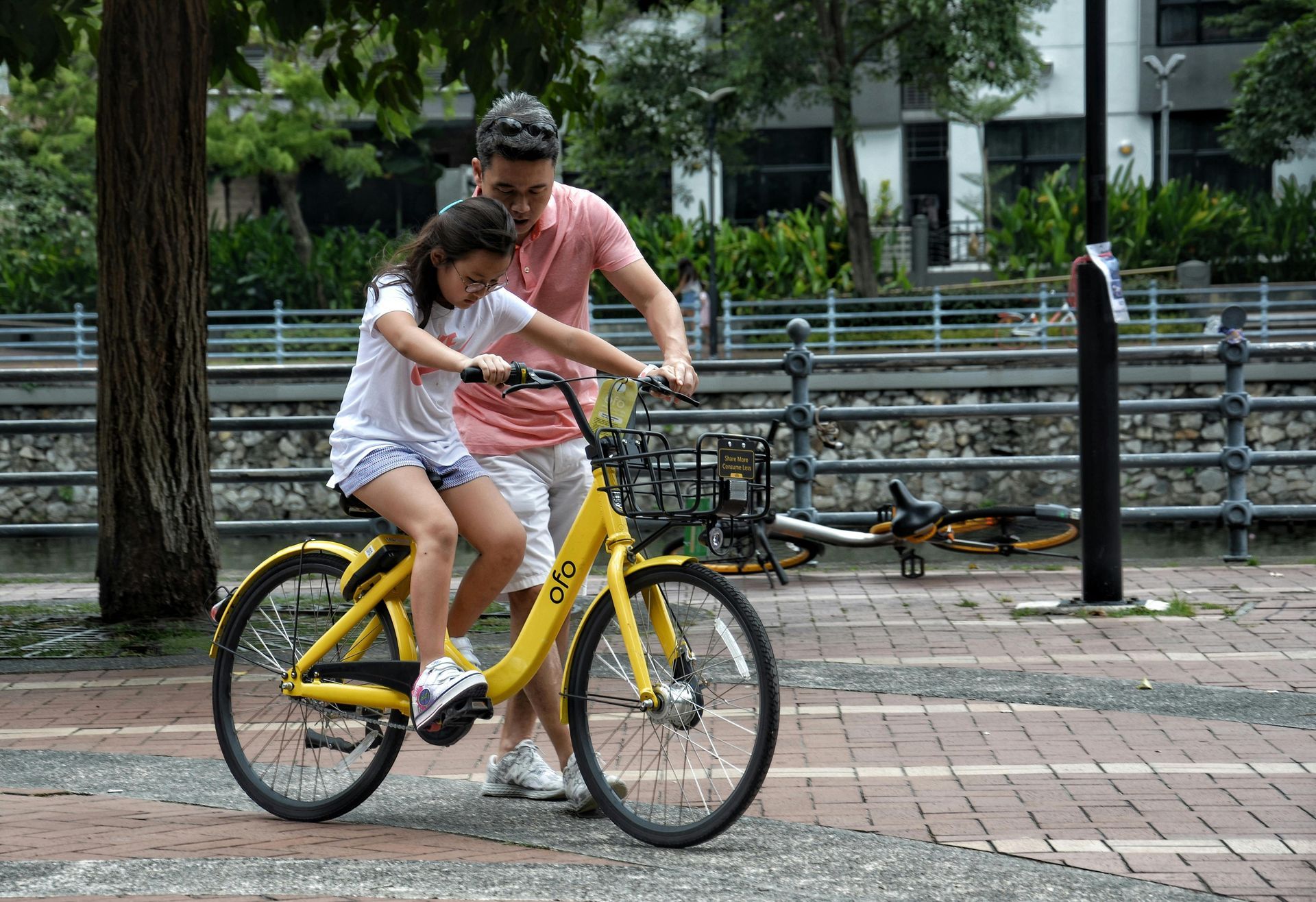 A man in a pink shirt helps a young girl learn to ride a yellow bicycle near a train track.