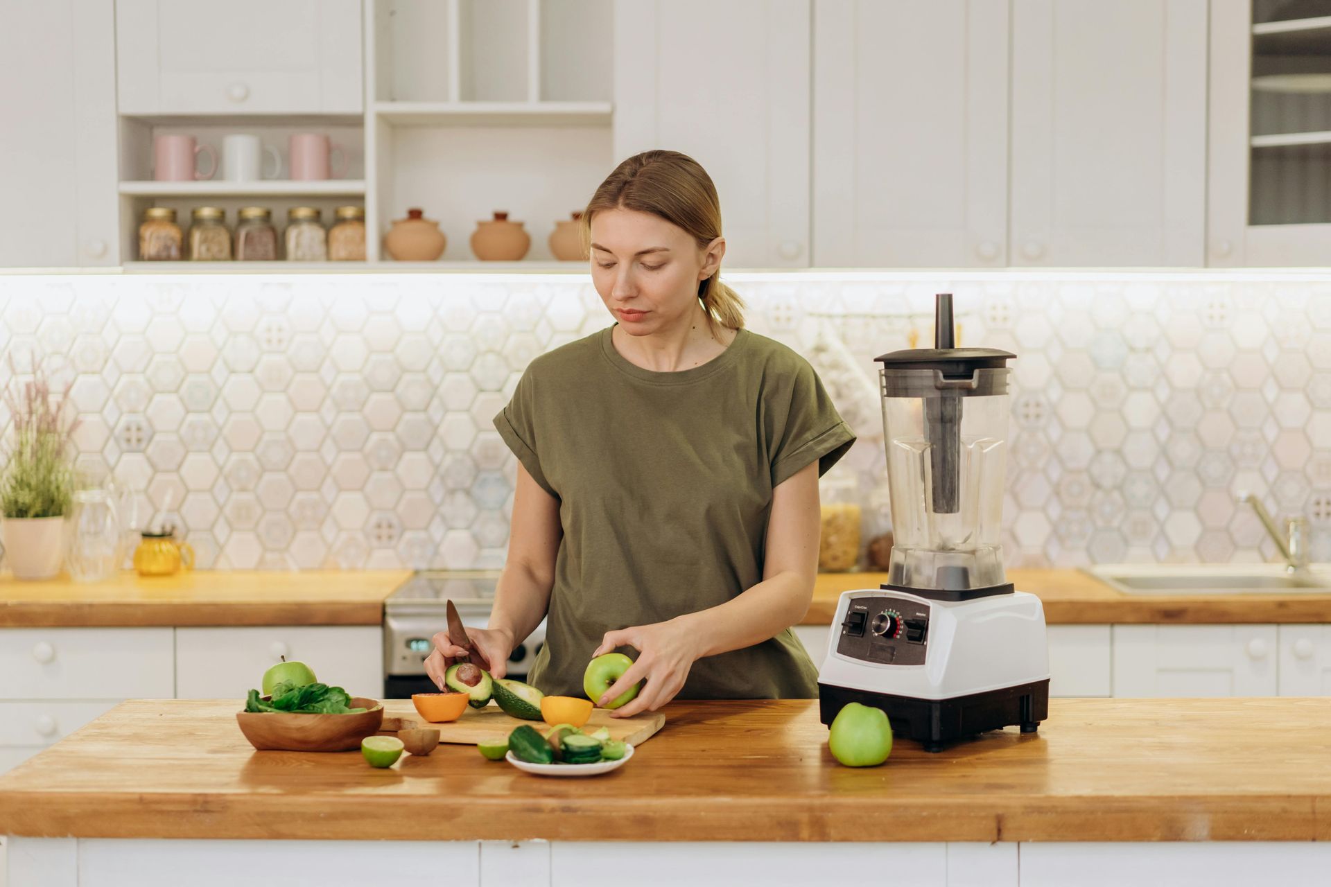 Woman in a green shirt prepping fruits and vegetables with a blender on a wooden countertop in a kitchen.