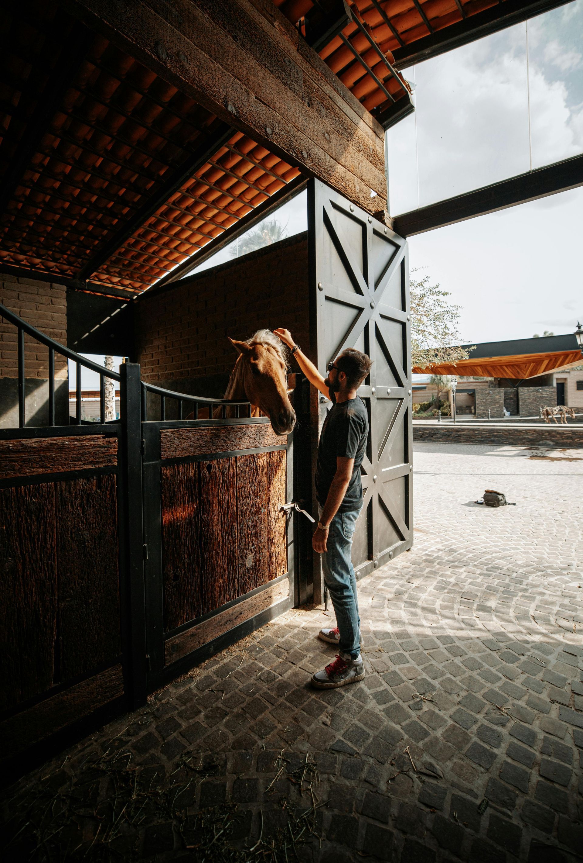 A person in a barn grooms a light brown horse. The setting is a brick-paved stable.