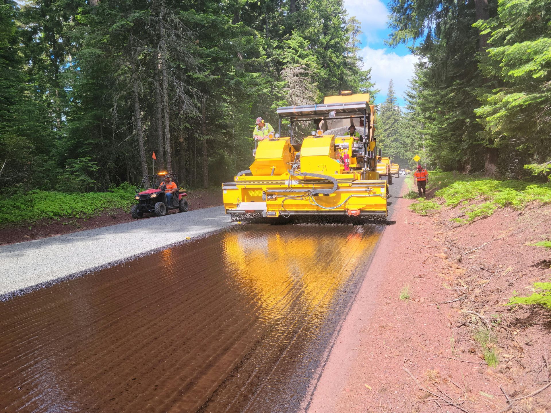 Road paving machine laying asphalt on a gravel road in a forest, with workers supervising.
