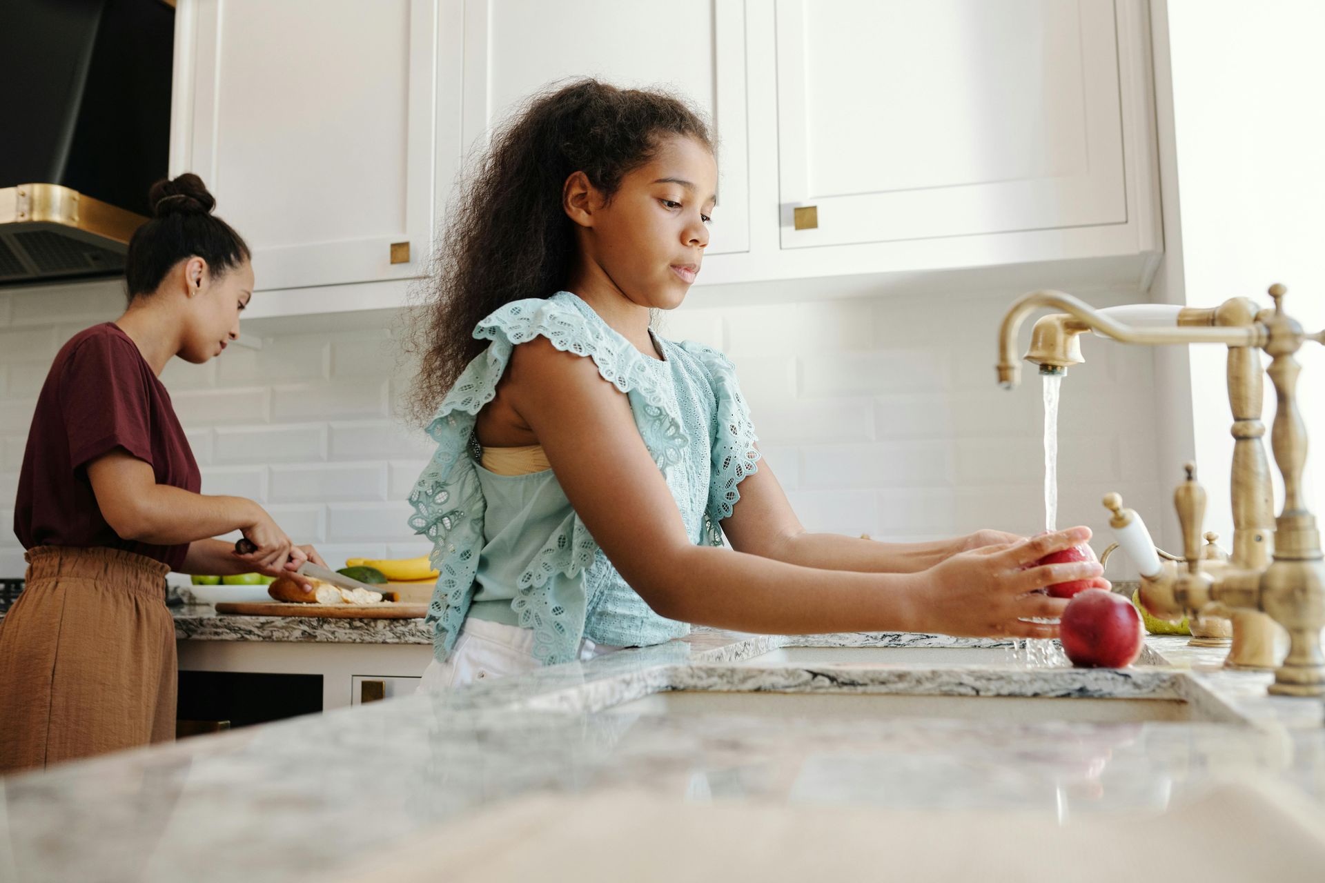 A young girl washing apples in a kitchen sink while her mother preps food. The kitchen is bright and white.