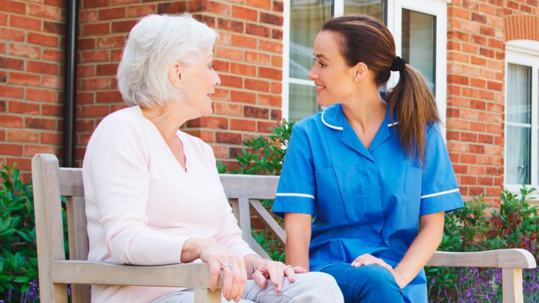 Two women sit on a bench, smiling and talking outside a brick building.