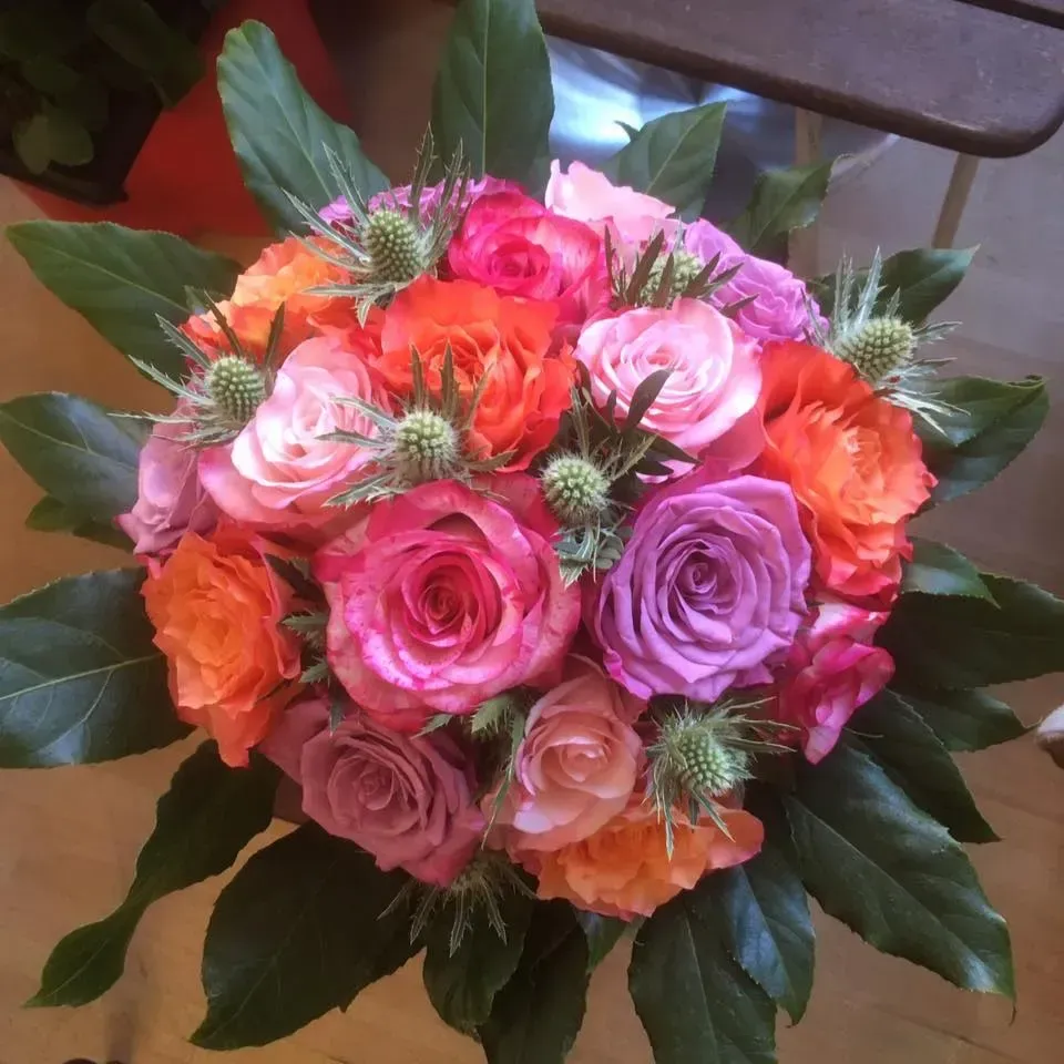 Buket af livlige orange, lyserøde og lilla roser med grøn tidsel og blade.