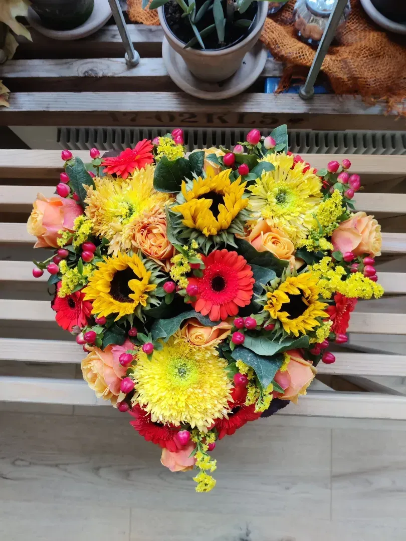 Hjerteformet blomsterarrangement med gule solsikker, røde gerberaer og orange roser, placeret på en træbænk.