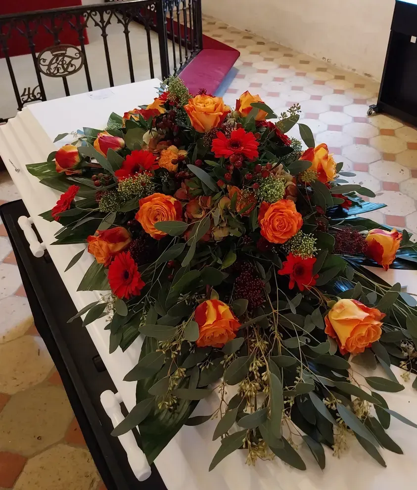 Blomsterarrangement af orange roser og grønt på en hvid kiste i en kirke.