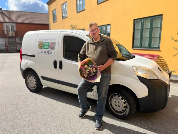 En mand med blomster står ved en hvid varevogn med et blomsterbutikslogo uden for en gul bygning.