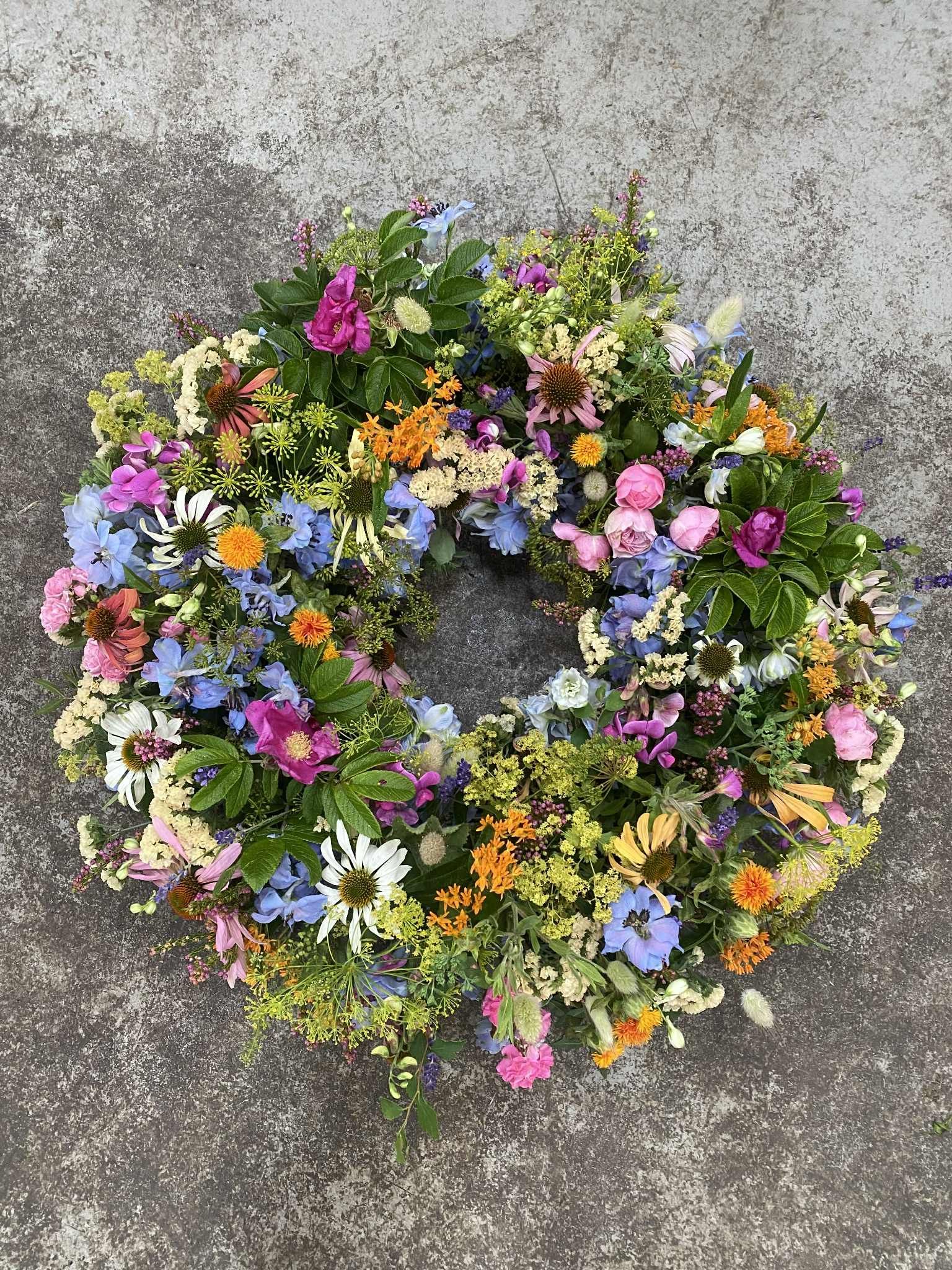 Farverig blomsterkrans på en grå betonoverflade med lyserøde, orange, blå og gule blomster.