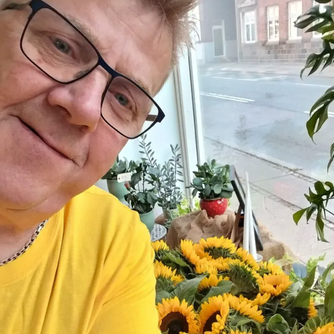 Mand i gul skjorte og briller smiler nær solsikker i et butiksvindue.