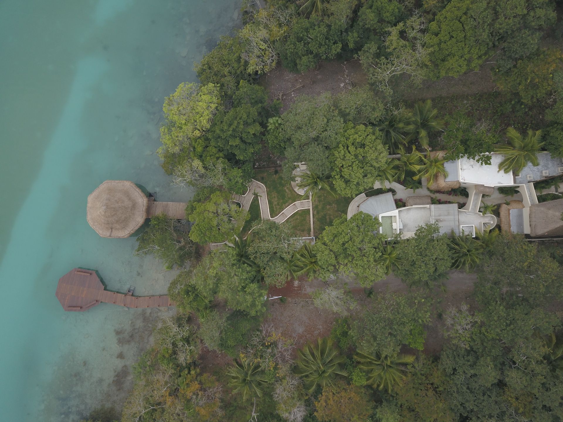 Aerial view of a structure on a shoreline. Turquoise water and a wooden dock with a thatched-roof shelter are visible.