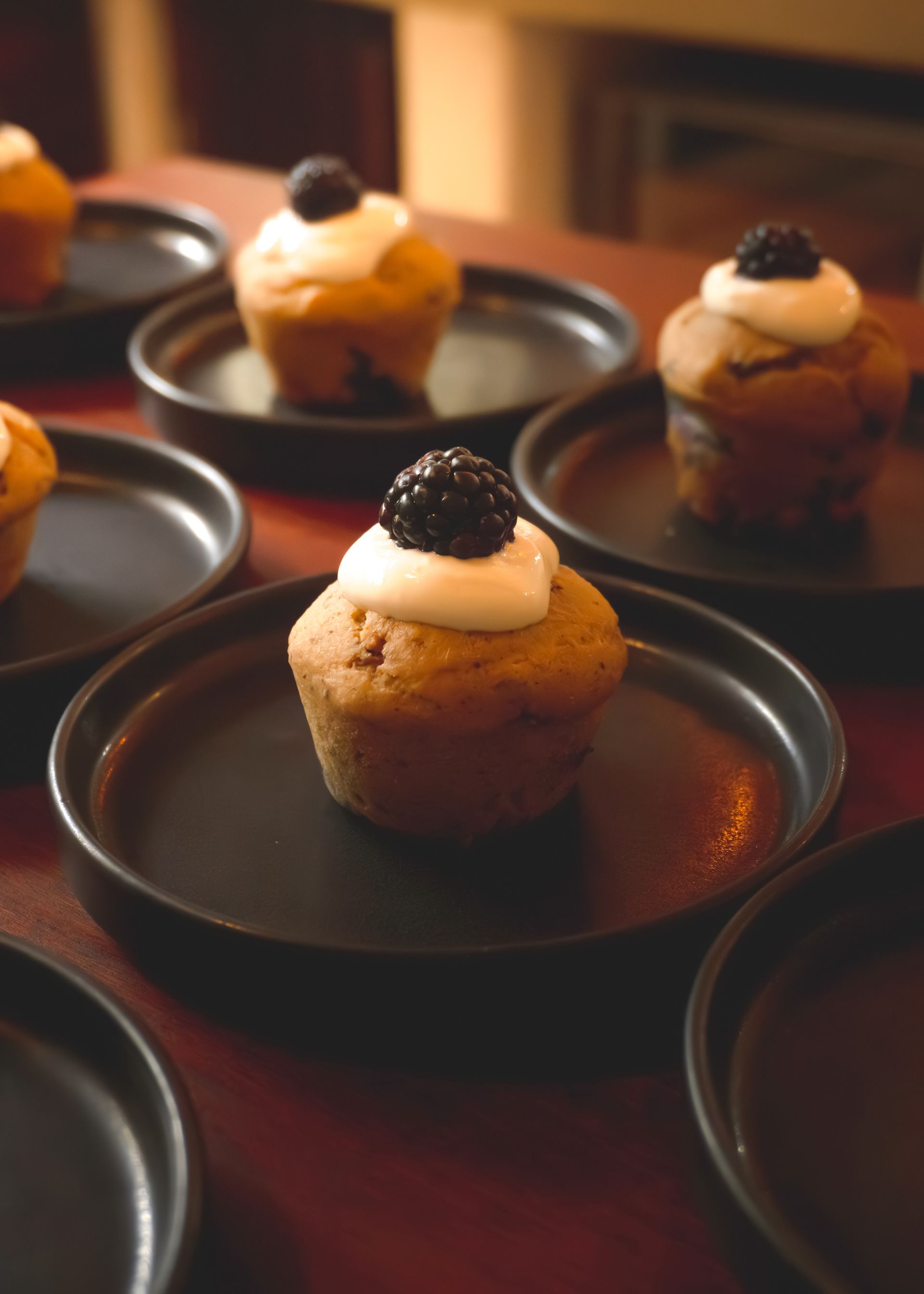 Muffins with white frosting and blackberries on black plates, arranged on a red table.