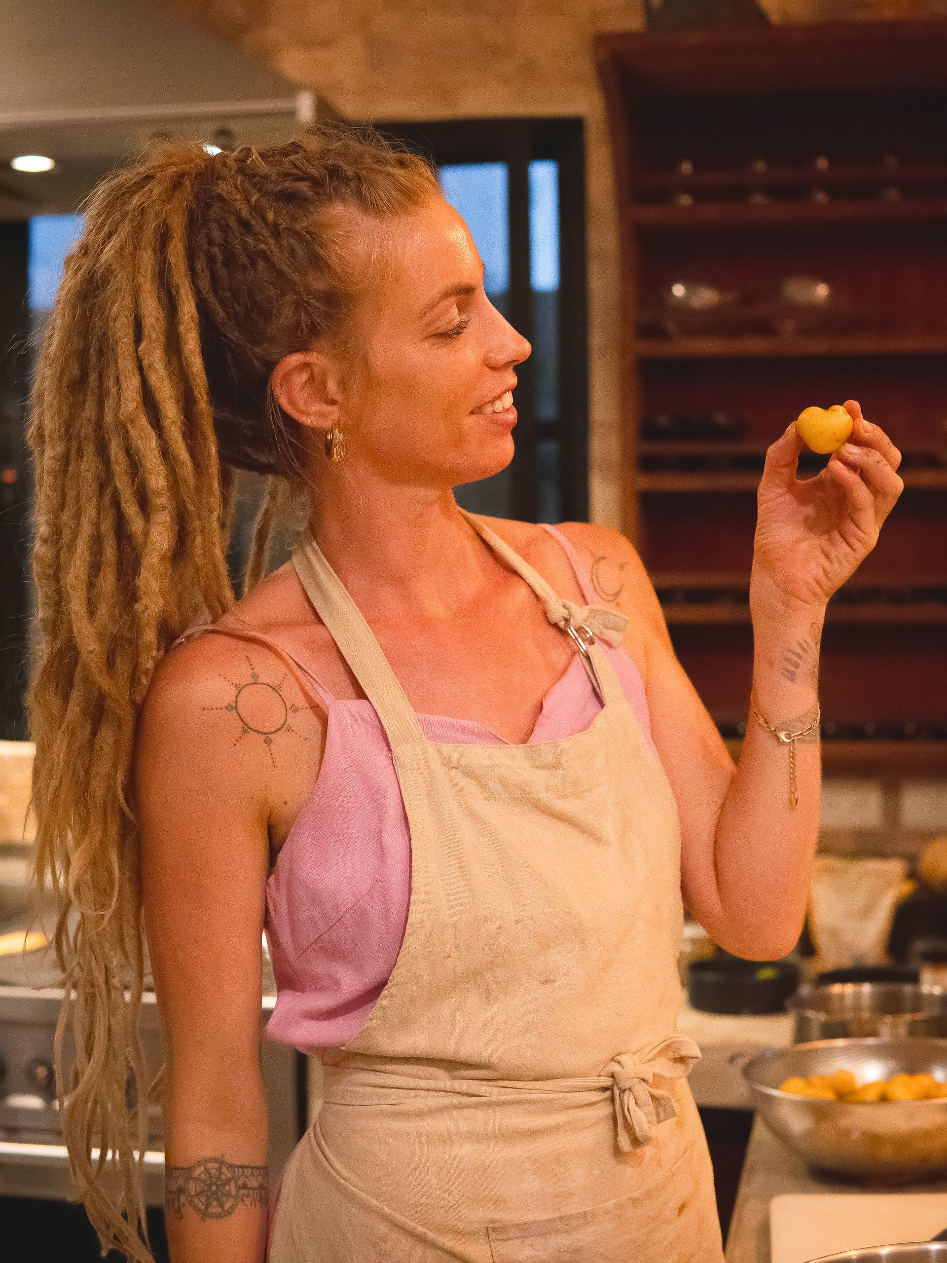 Woman with blonde dreadlocks holding a yellow fruit, wearing a pink top and apron in a kitchen.