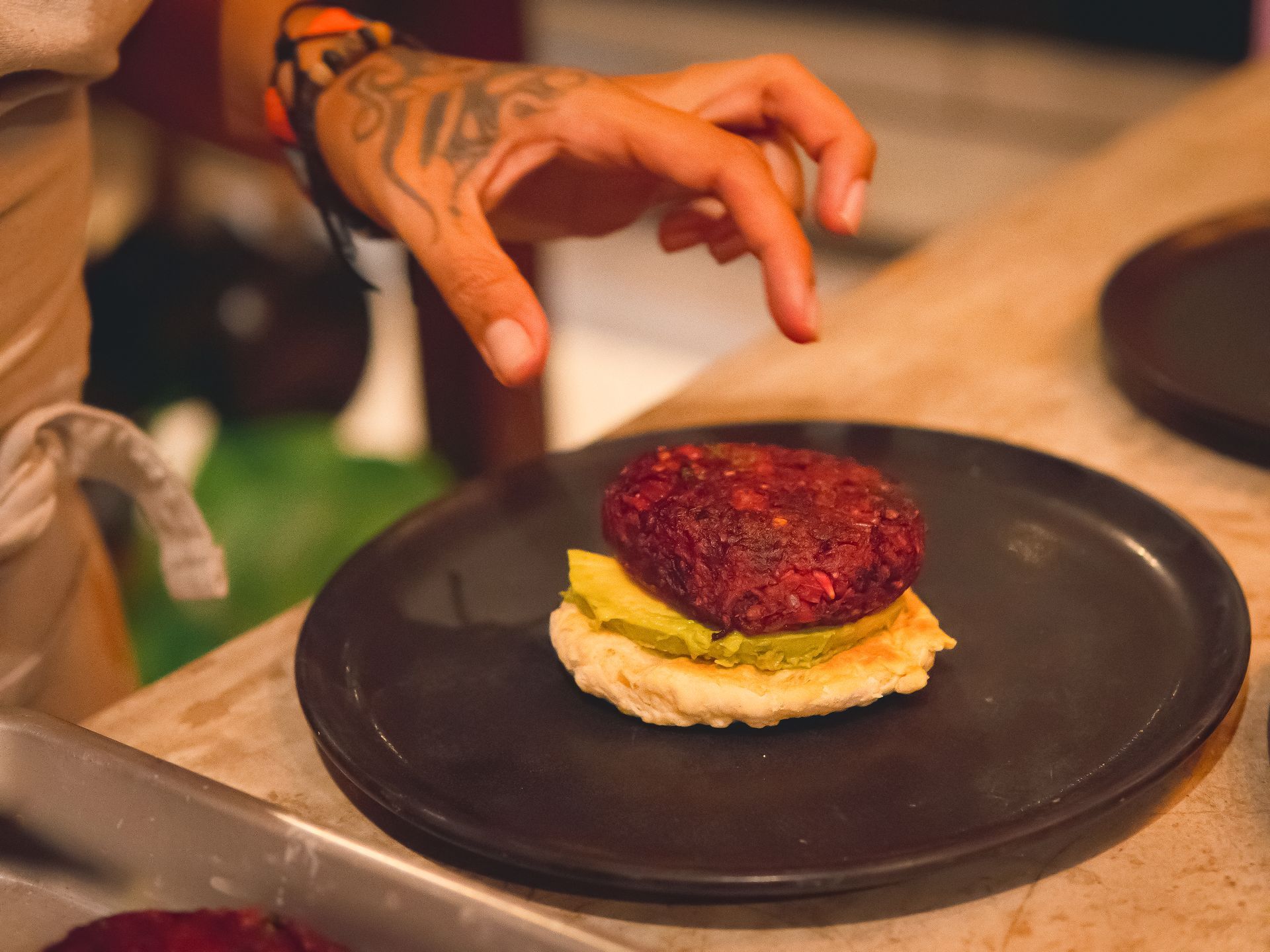 Chef garnishing a plant-based burger. Beet patty on avocado spread, atop a bun, on a black plate.