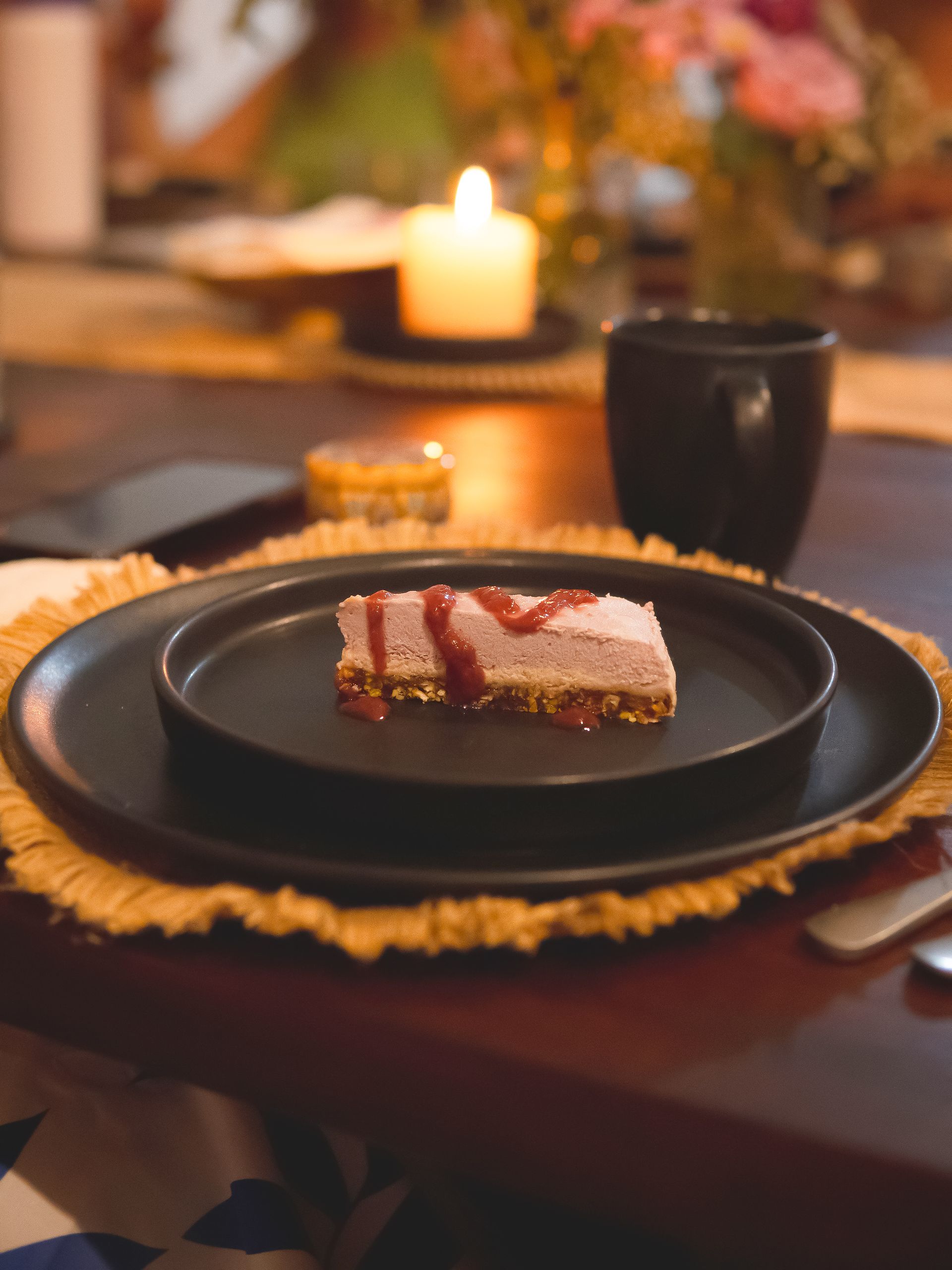 Dessert slice on black plate, drizzled with sauce, on a table set with candles and a mug.