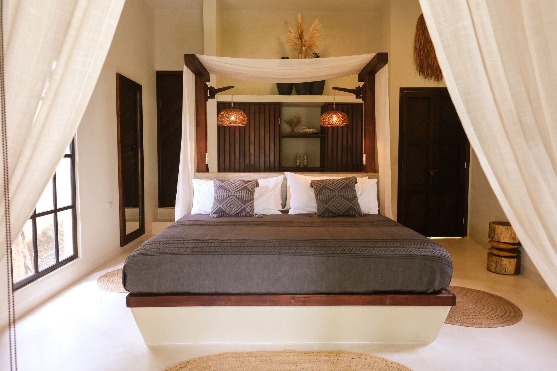 Bedroom with canopy bed, neutral tones, wooden accents, sheer curtains, and woven rugs.