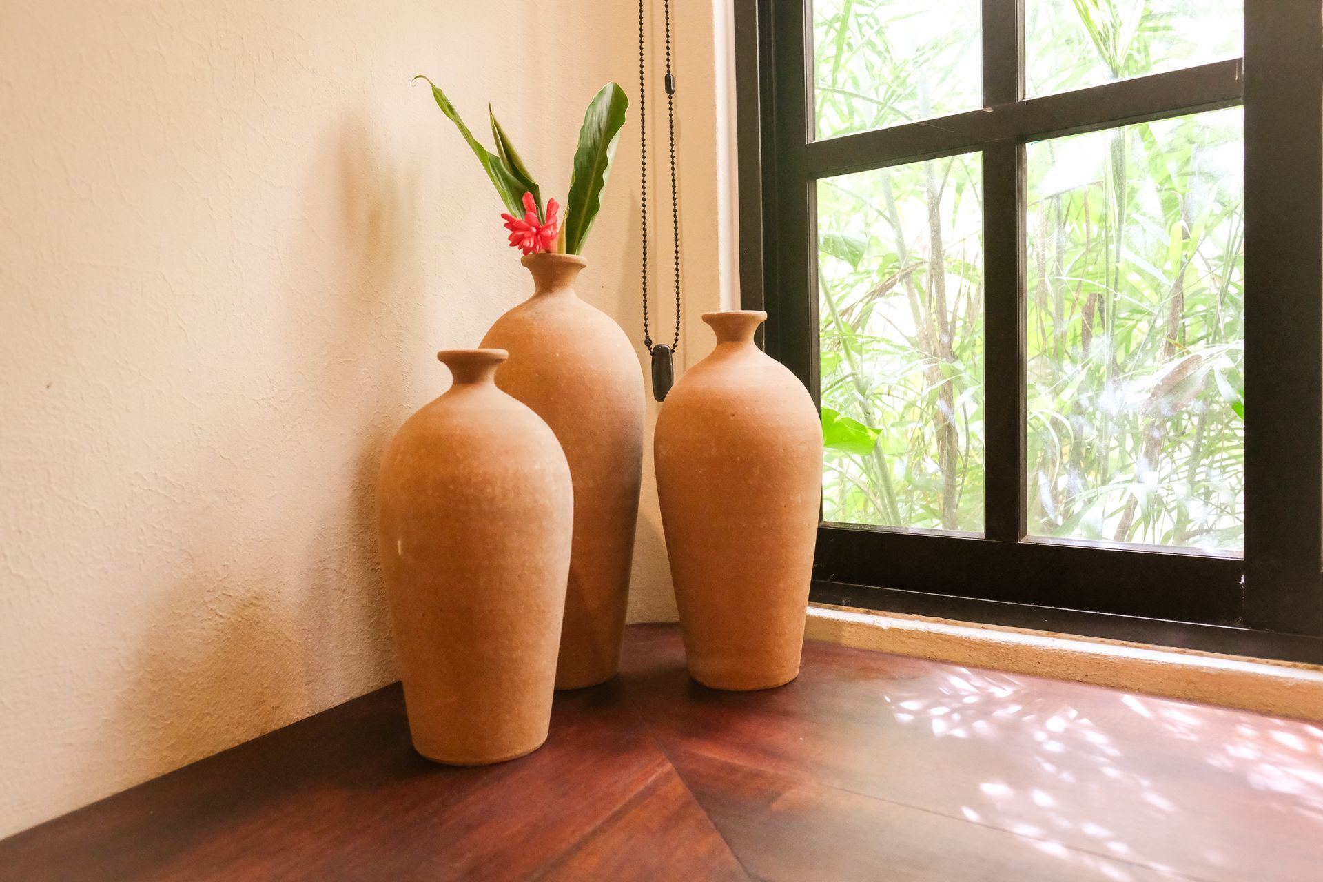 Three brown vases on a wooden floor next to a window, one vase with a red flower and green leaves.