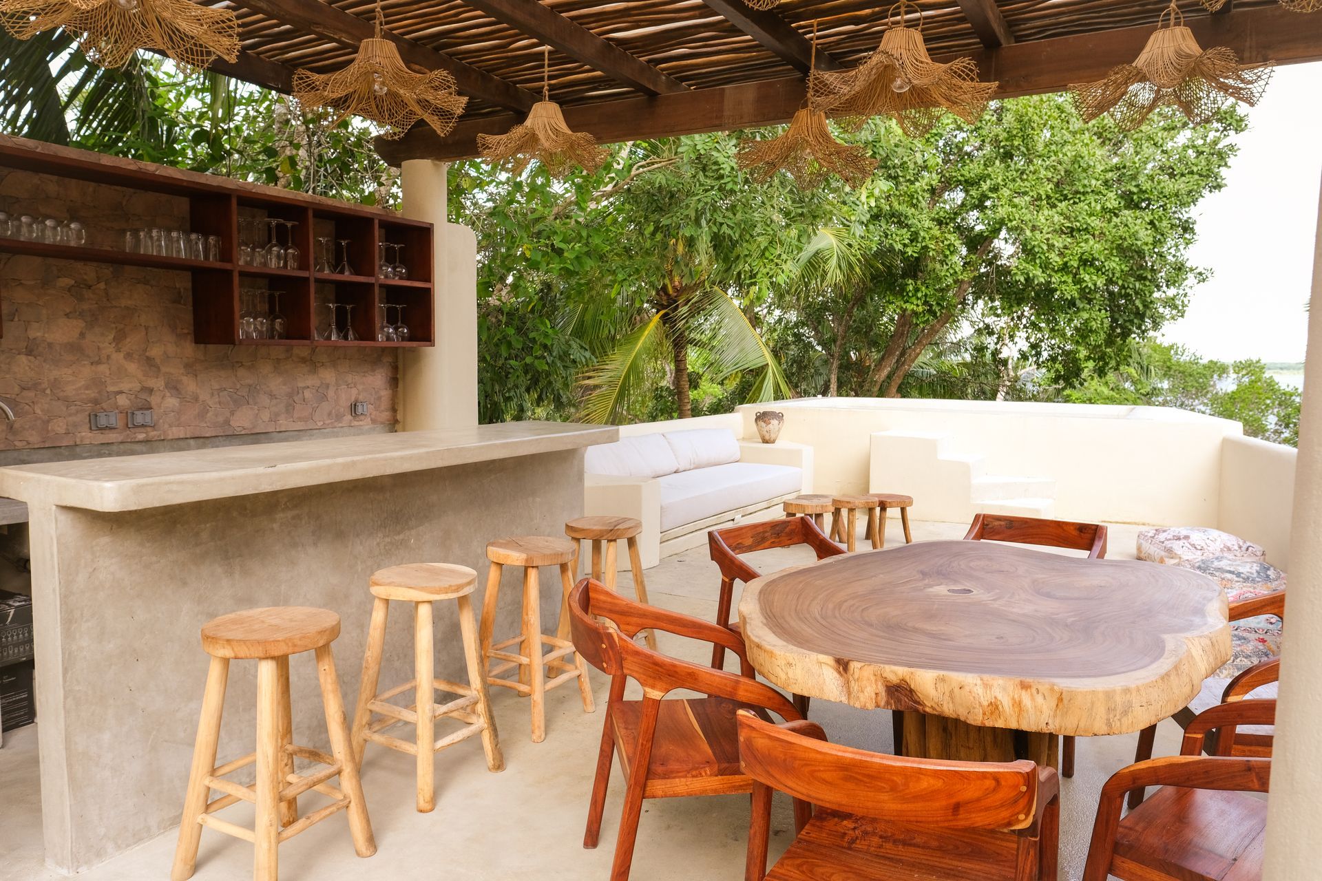 Outdoor bar with wooden stools, a round table, and chairs; trees in the background.