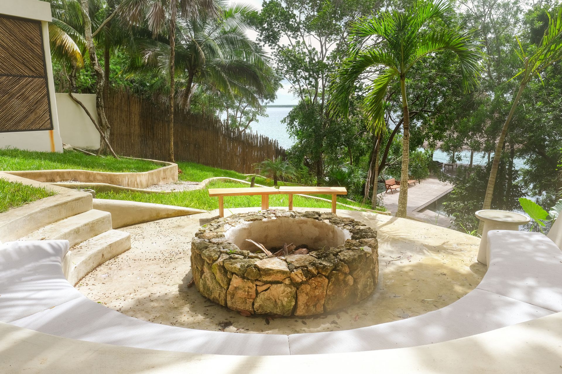 Fire pit with seating in a tropical outdoor setting. Grass, trees, and ocean views.