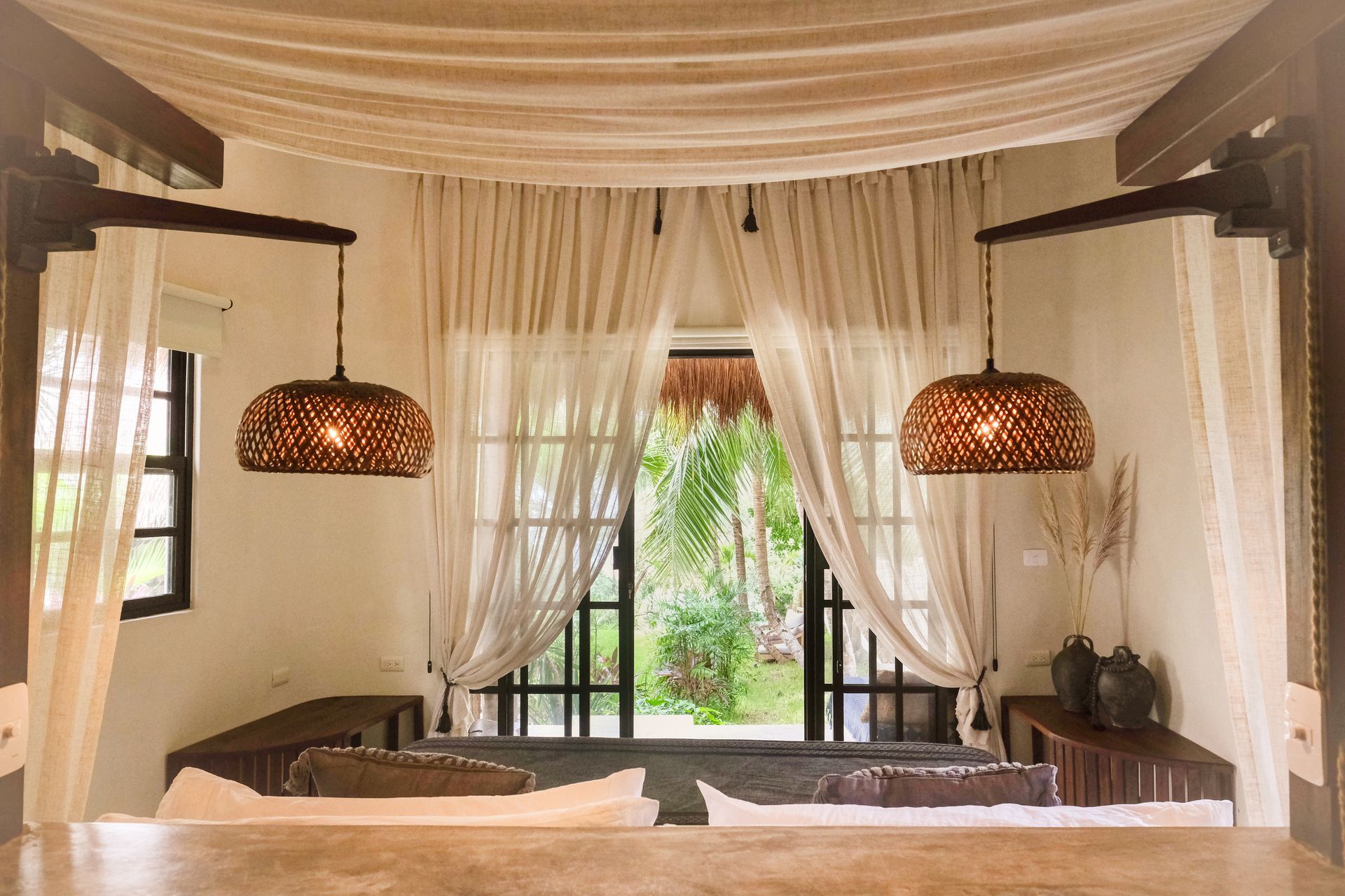 Bedroom with woven pendant lights, sheer curtains, and open doors to a lush garden.