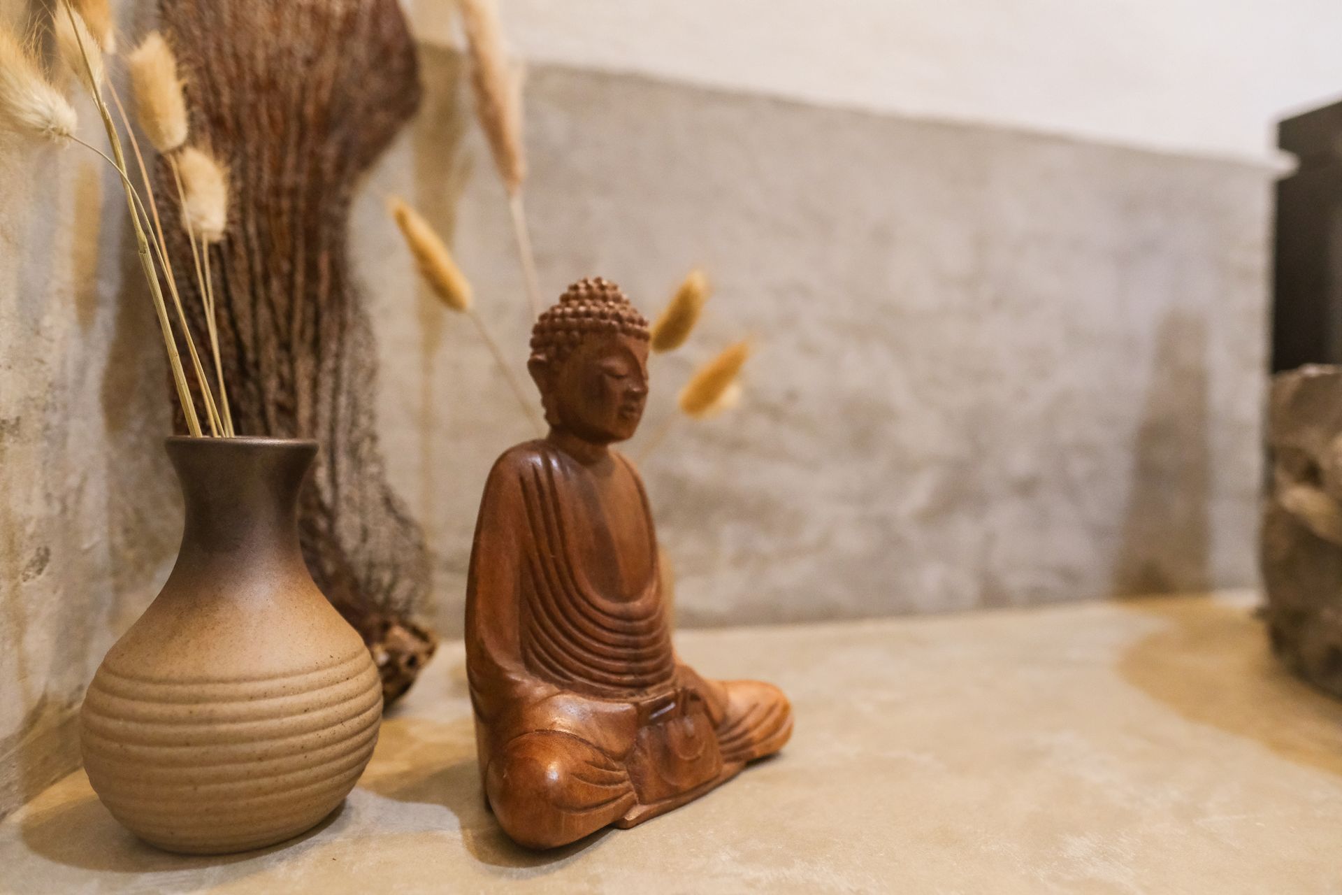 Wooden Buddha statue, seated in meditation, next to a vase with dried flowers, on a concrete surface.