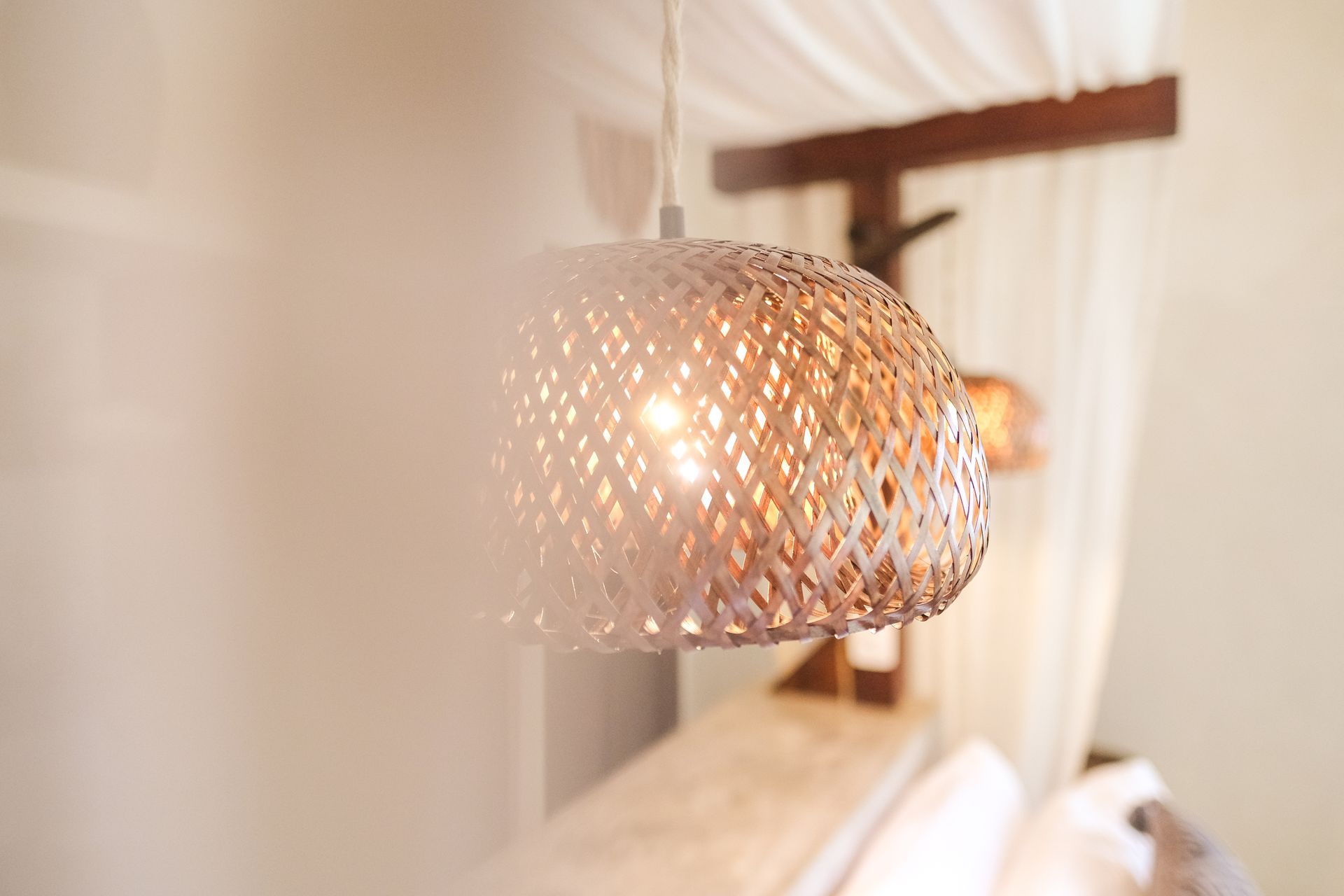 Woven pendant lamp with warm light, hanging above a bed with white bedding, soft focus.