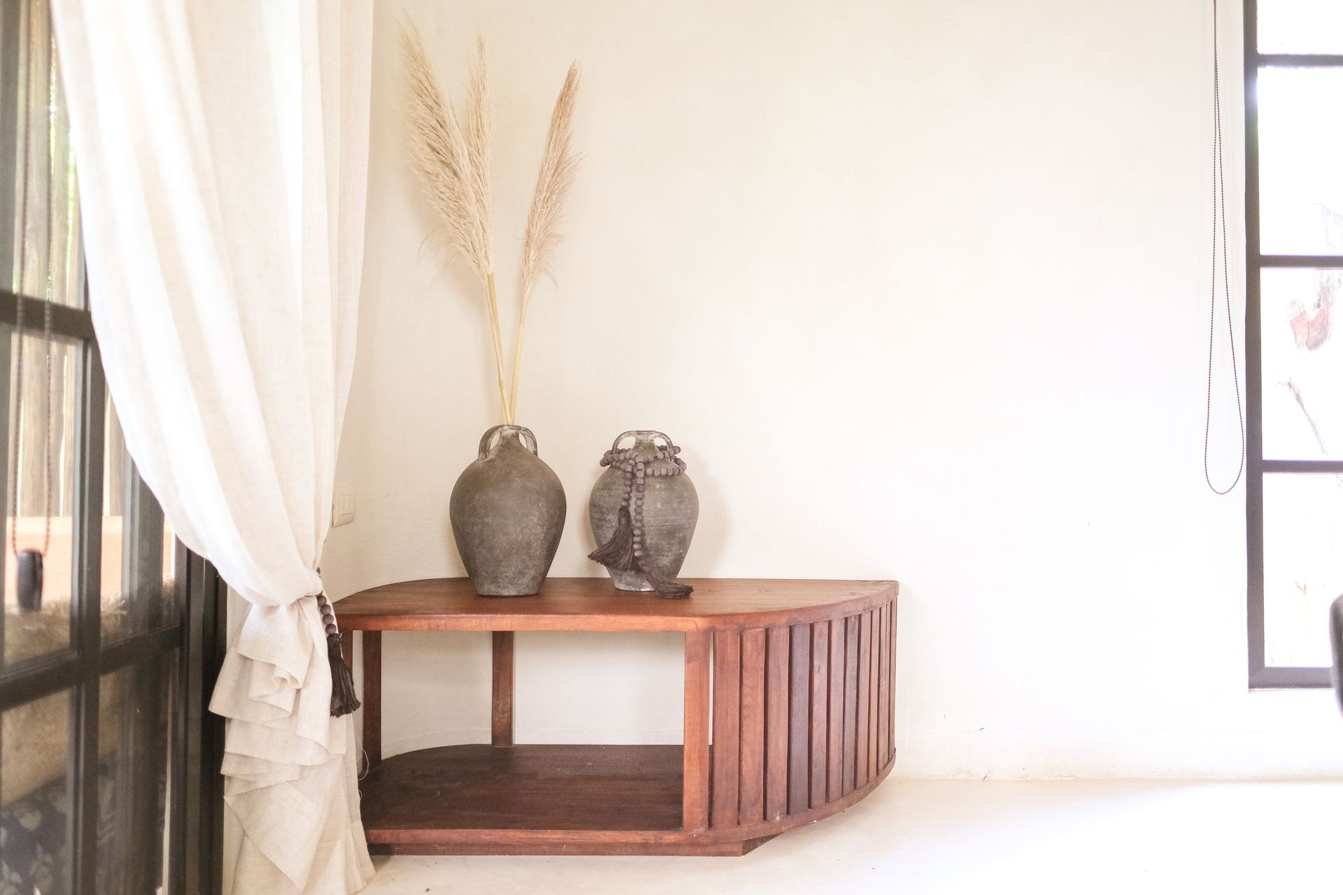 Wooden table with two vases and dried plants against a white wall, near a window and curtain.