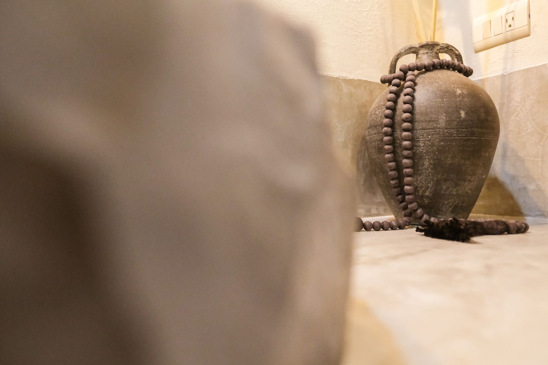 Large, dark vase with beaded necklace and reed diffuser against a neutral wall.