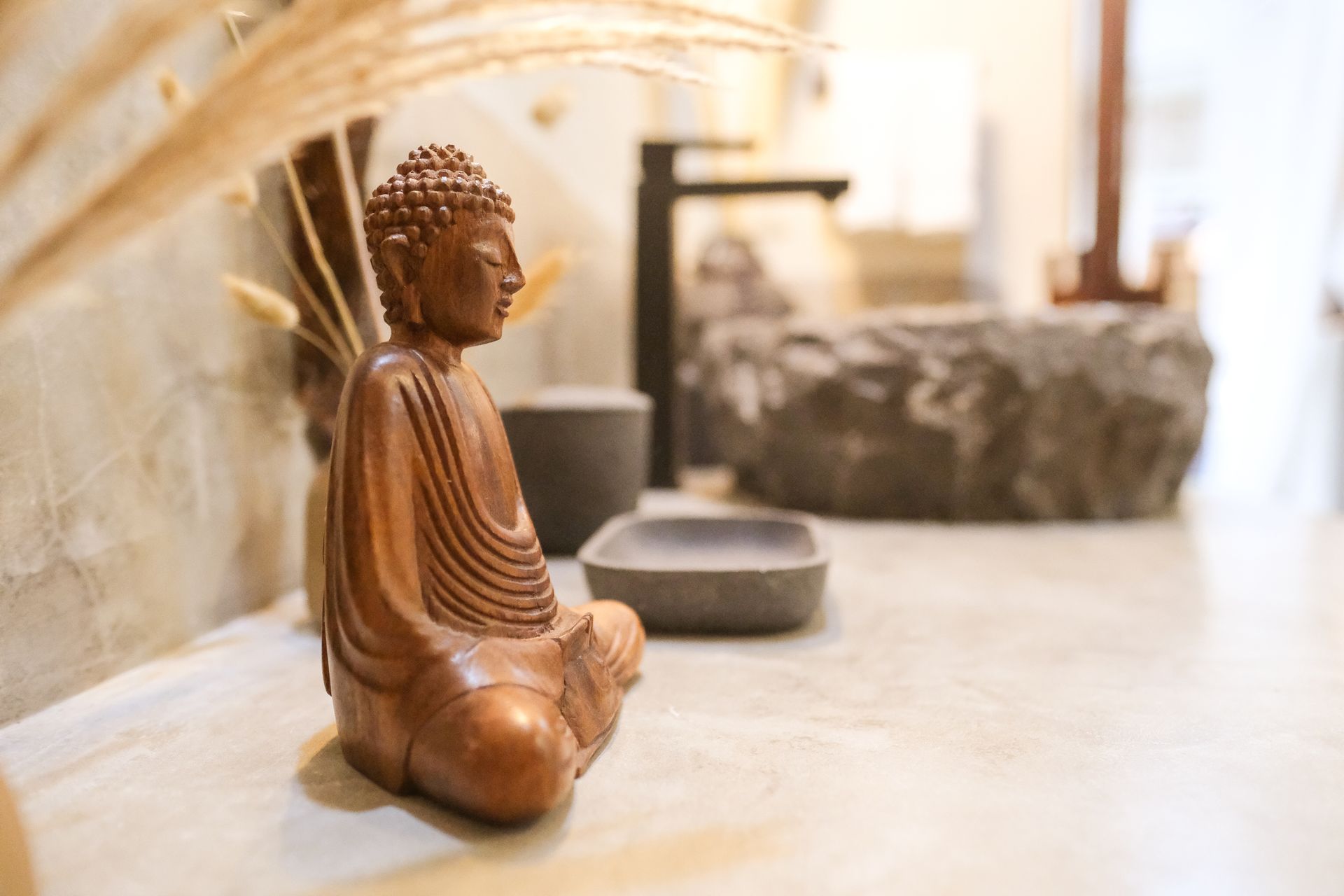 Wooden Buddha statue in a meditative pose, with a blurred background of a bathroom sink and decor.