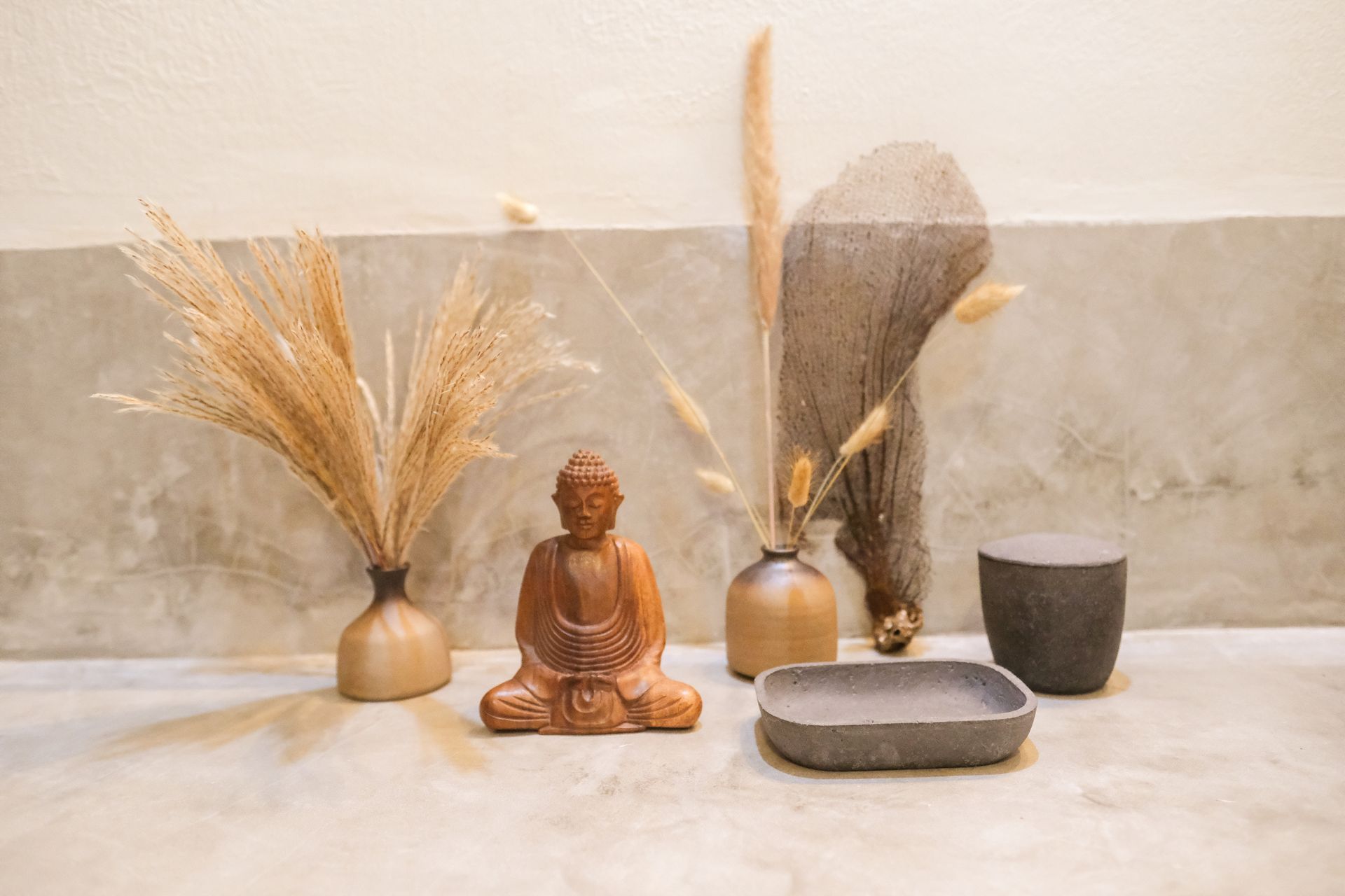 Wooden Buddha statue with vase arrangements and a stone dish against a neutral-toned wall.