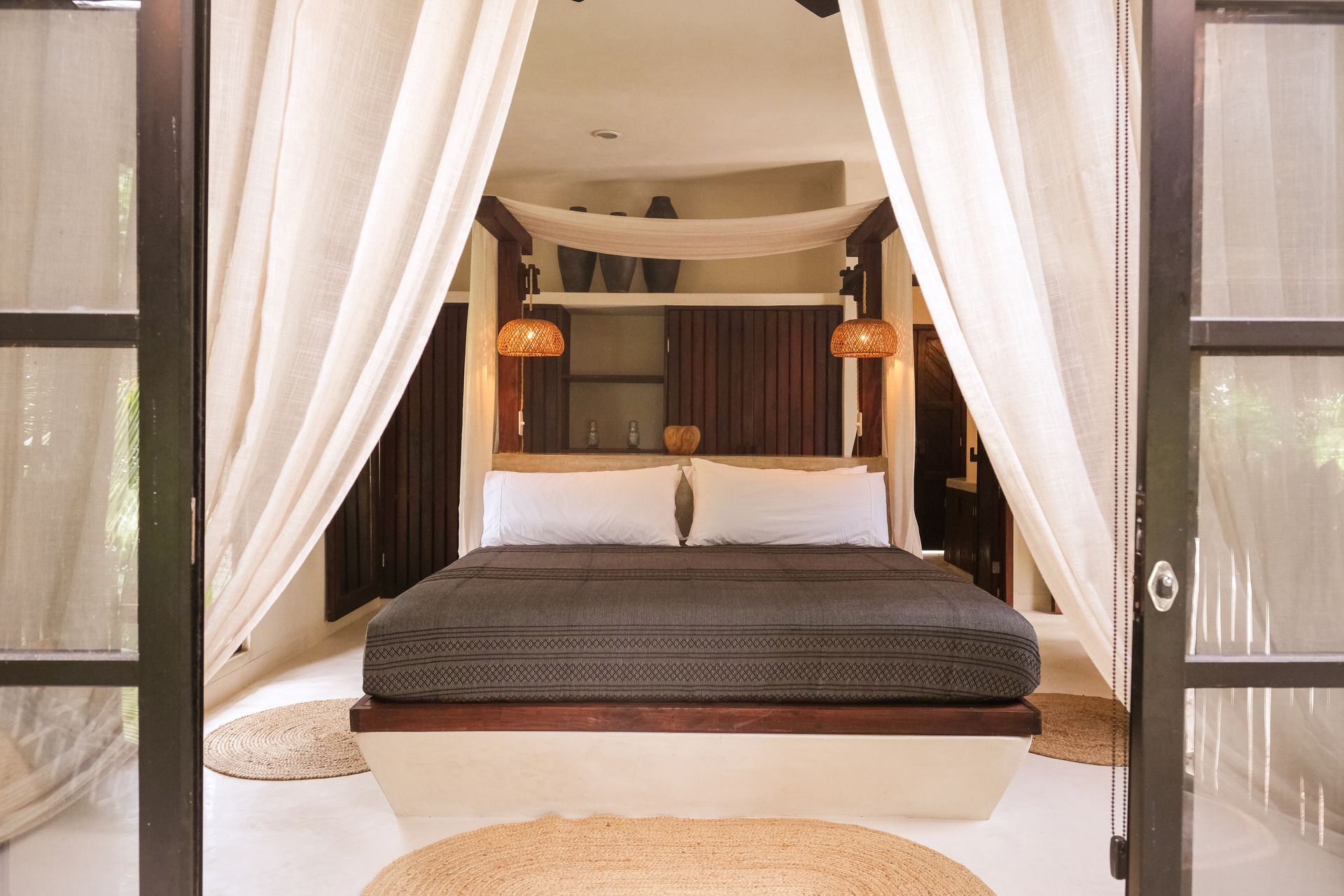Bedroom with canopy bed, dark bedding, and white curtains, viewed through open glass doors.