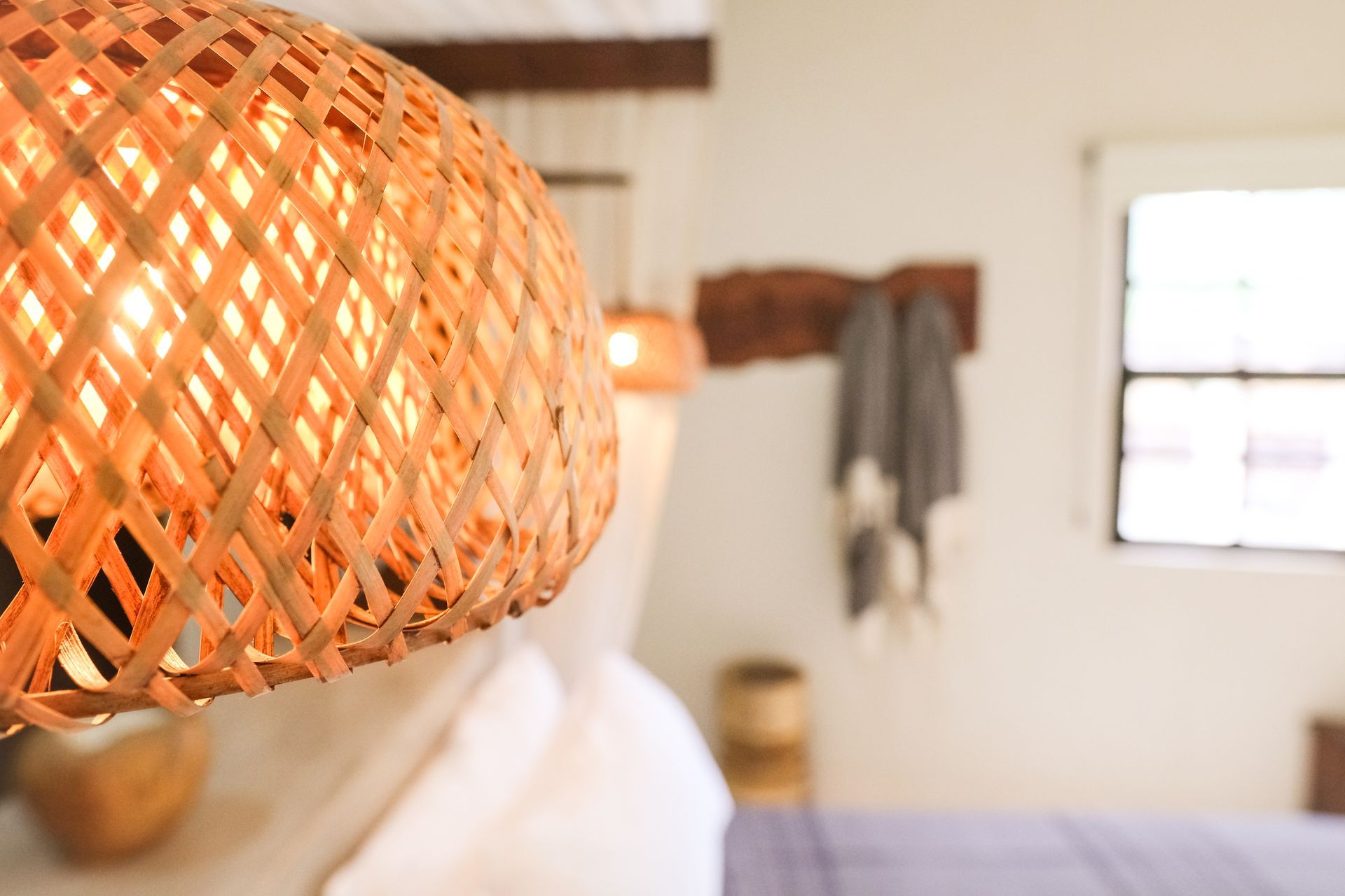 Woven light fixture hangs in a bedroom. The focus is on the fixture with a bed and window blurred in the background.