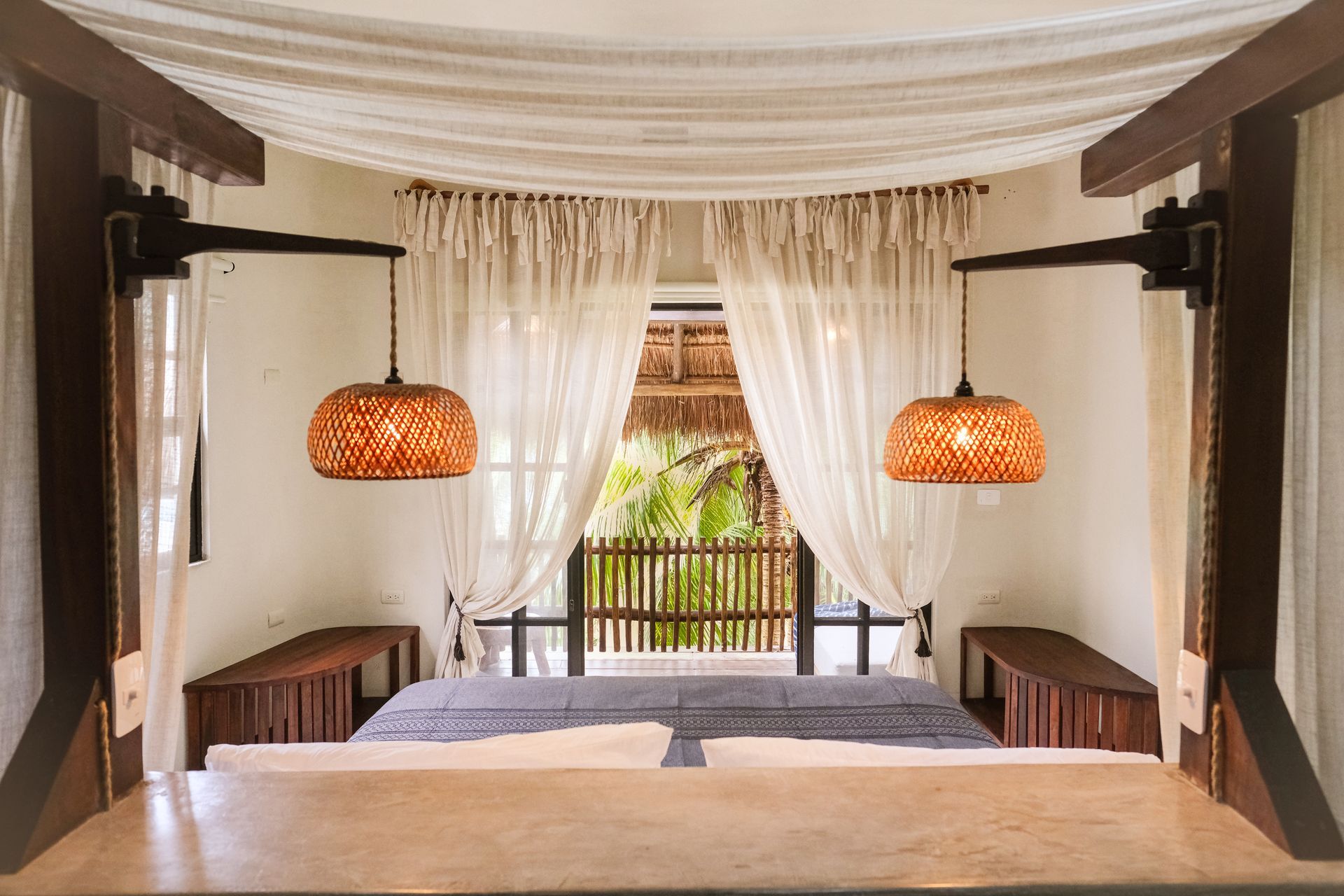 Bedroom with canopy bed, woven lamps, sheer curtains, and balcony view.