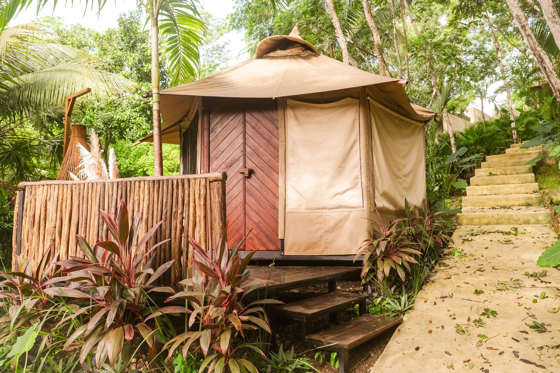 Small cabin with a thatched roof and wooden door, set in lush greenery, with stone stairs leading up to it.
