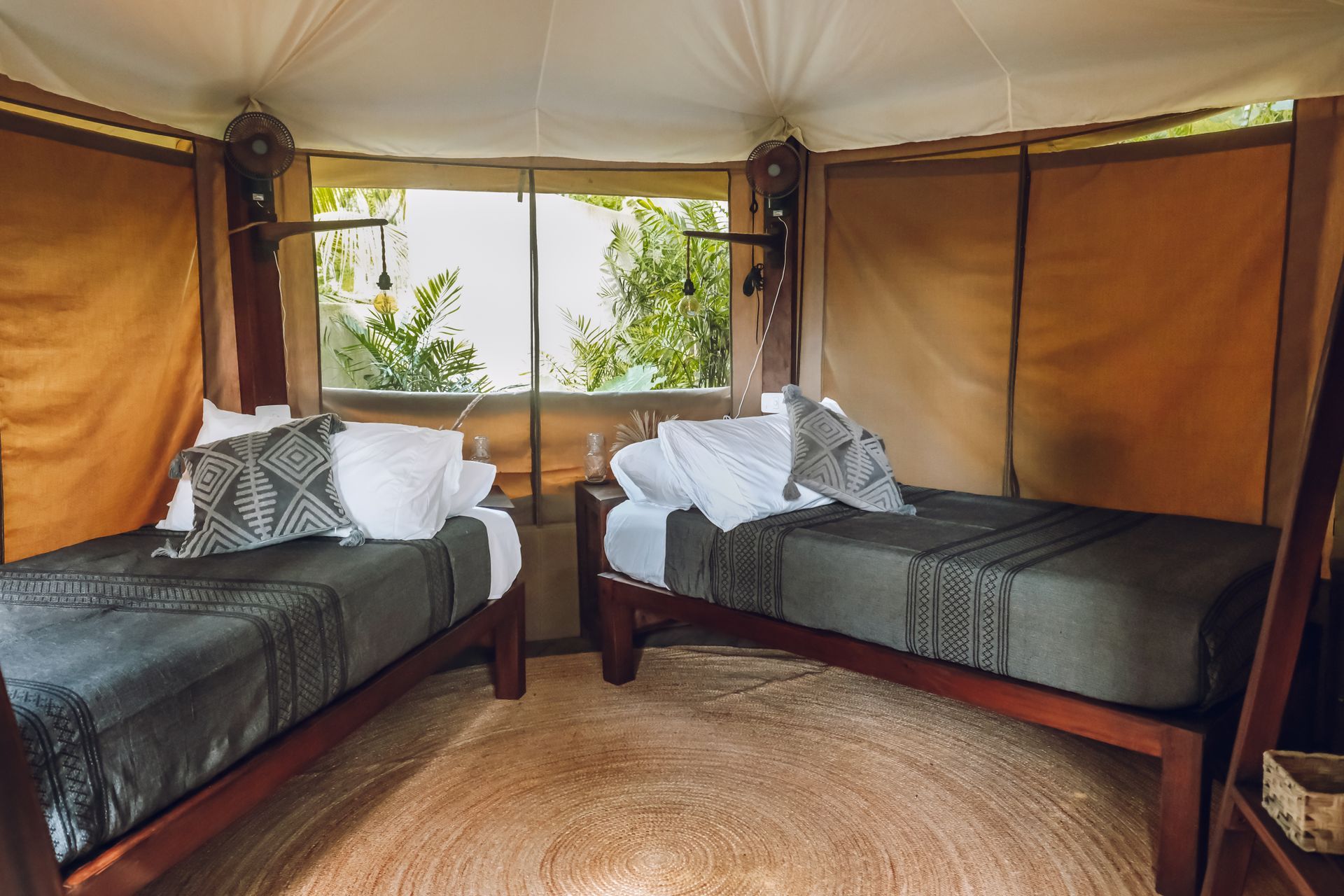 Two beds inside a tent with open window looking out at greenery. Brown wood frames, gray bedding, neutral tones.