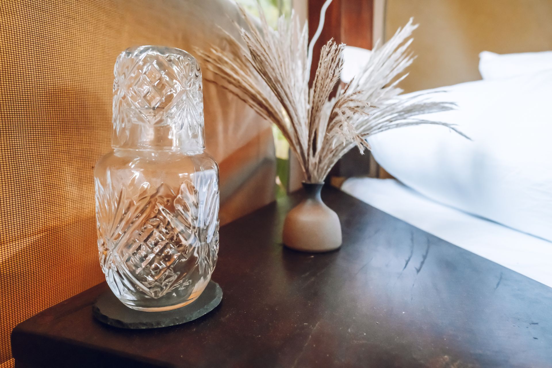 Cut-glass water carafe and glass, and dried ornamental grass in vase on a wooden nightstand.