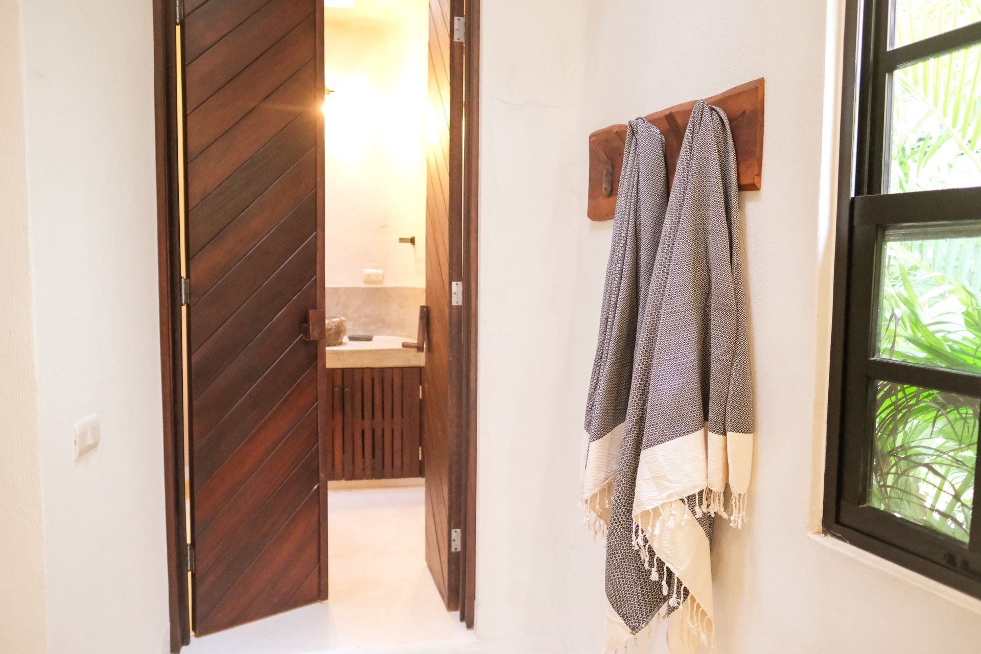 Wooden door opens to a bathroom, towels hang on wall, window to the right with greenery.